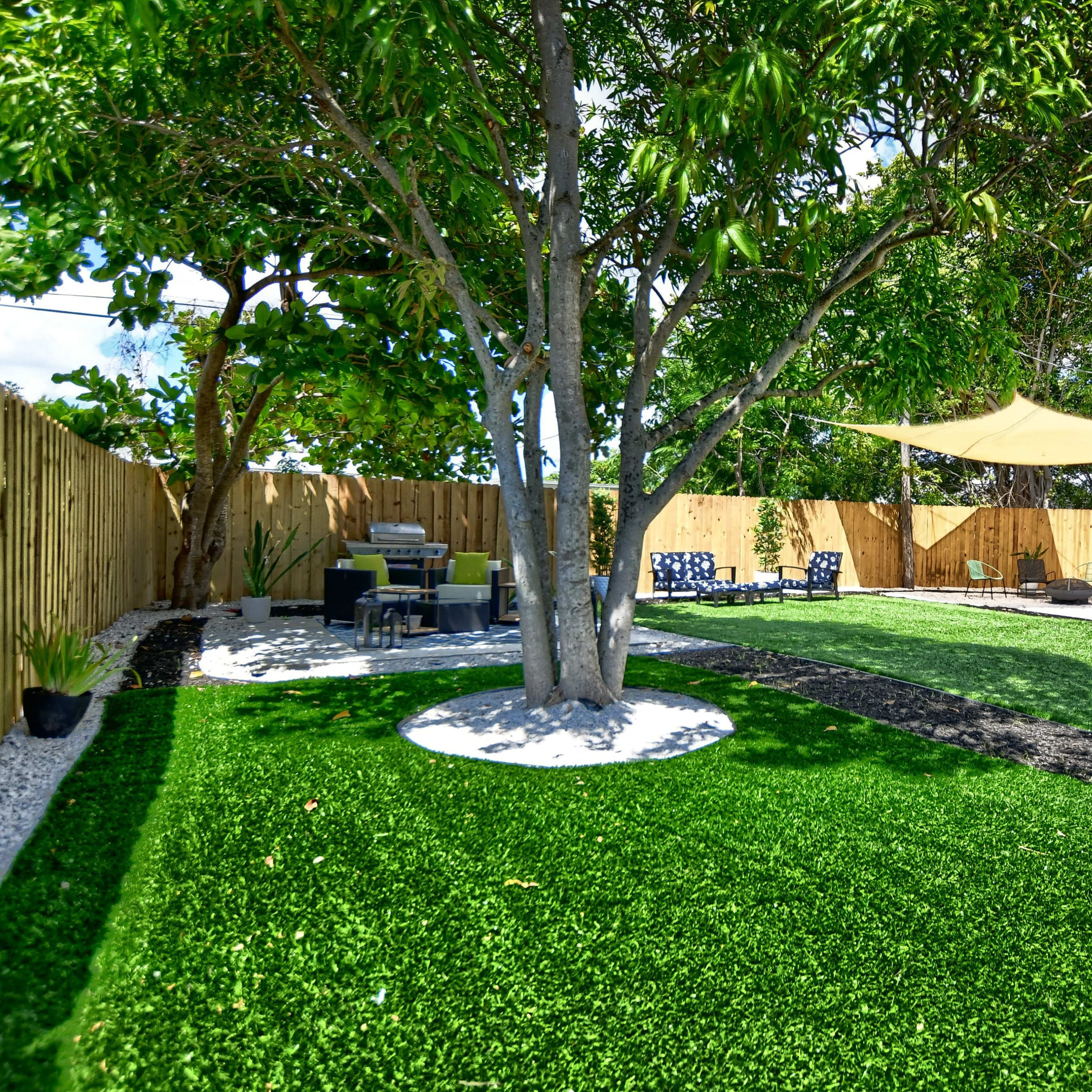 A landscaped backyard with artificial grass, a seating area under a tree, and a shaded structure to the side surrounded by a wooden fence.