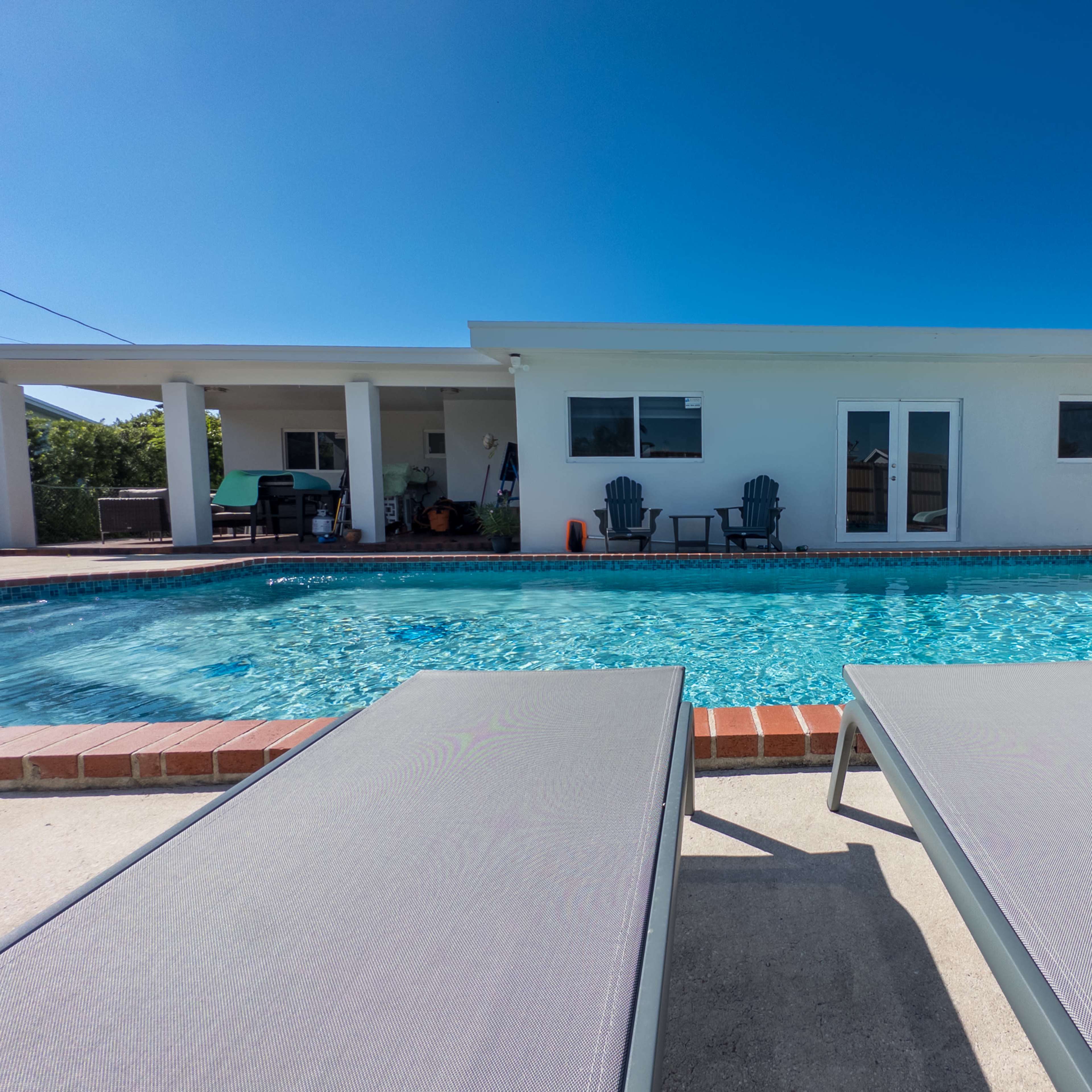 A swimming pool with lounge chairs is in front of a white house under a clear blue sky.