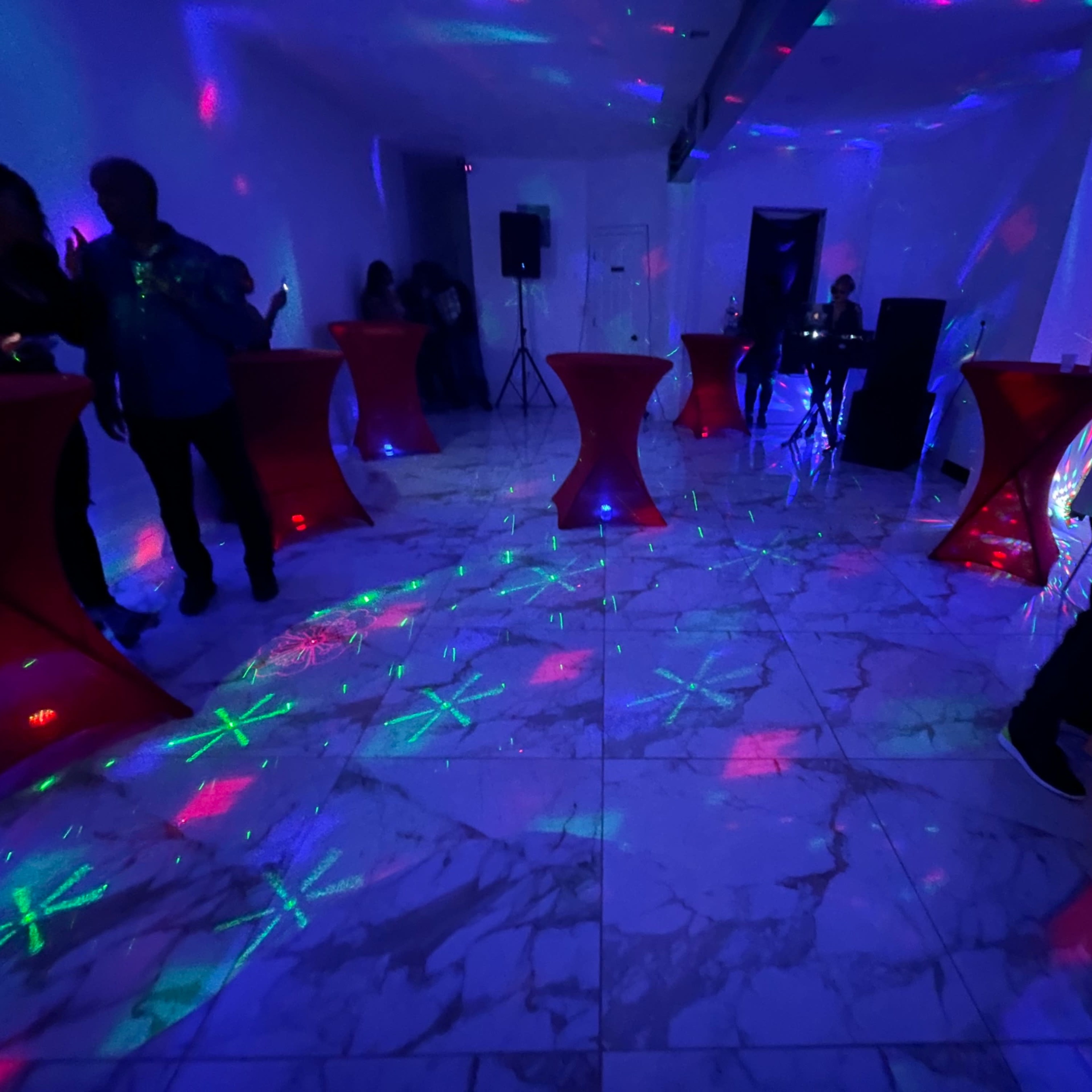 A dance floor with illuminated tables, colorful light projections, and people mingling in a brightly lit space.