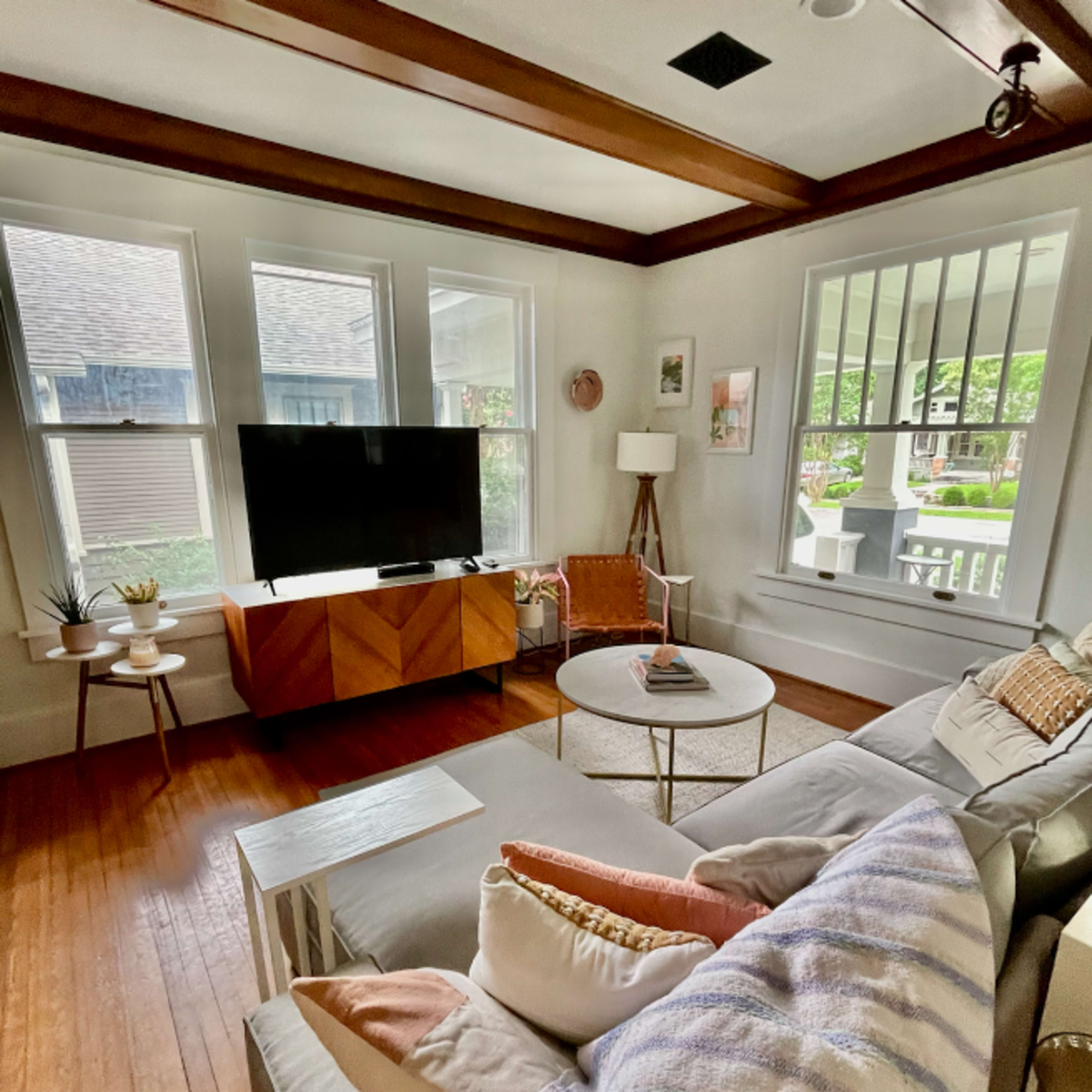The living room features a sectional sofa with decorative pillows, a coffee table, a wooden media cabinet holding a television, and large windows providing natural light.