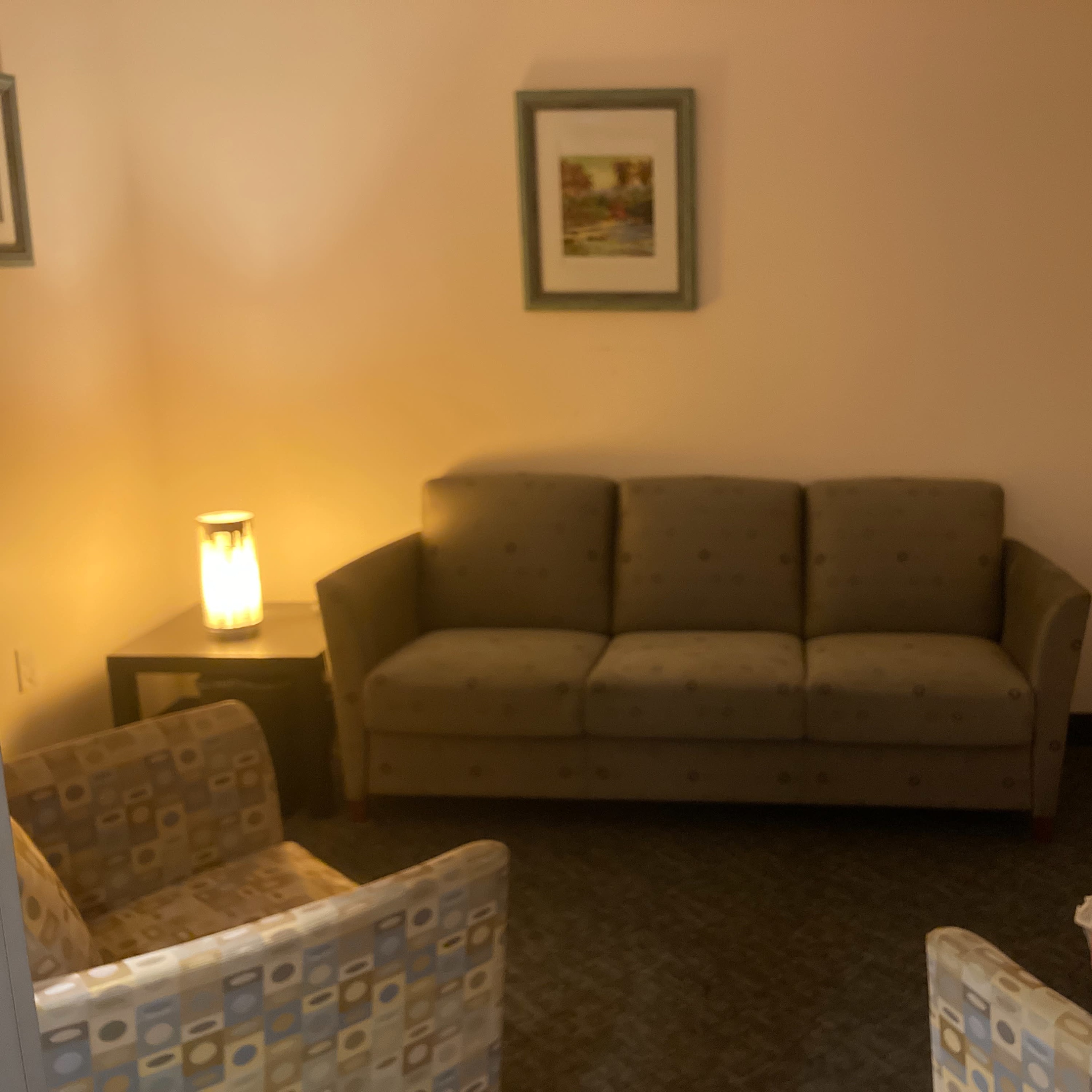 A small seating area with a gray couch and two patterned chairs arranged around a side table, illuminated by a lamp.