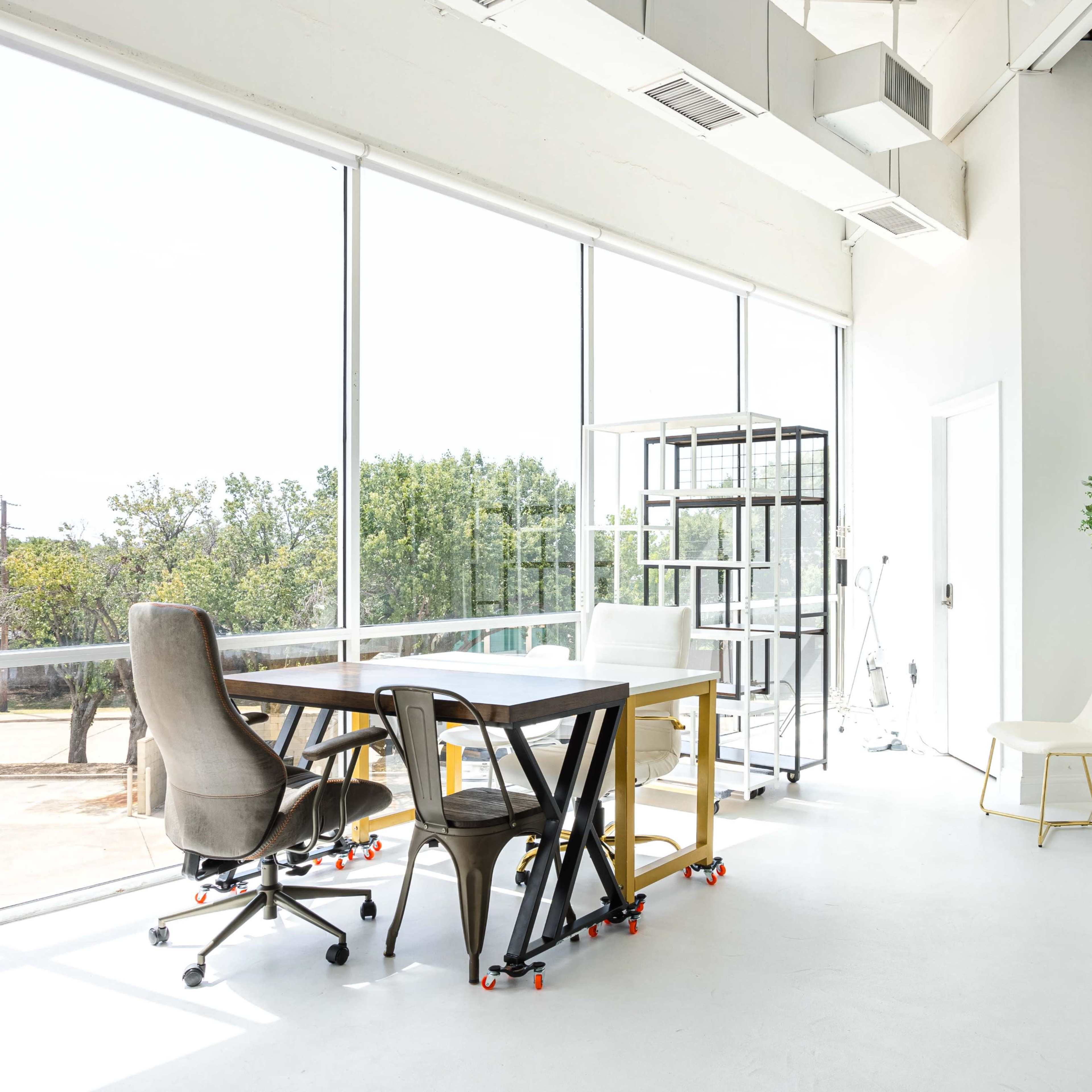 A bright, modern office space featuring large windows, a white floor, a black table with rolling chairs, and minimalistic decor.
