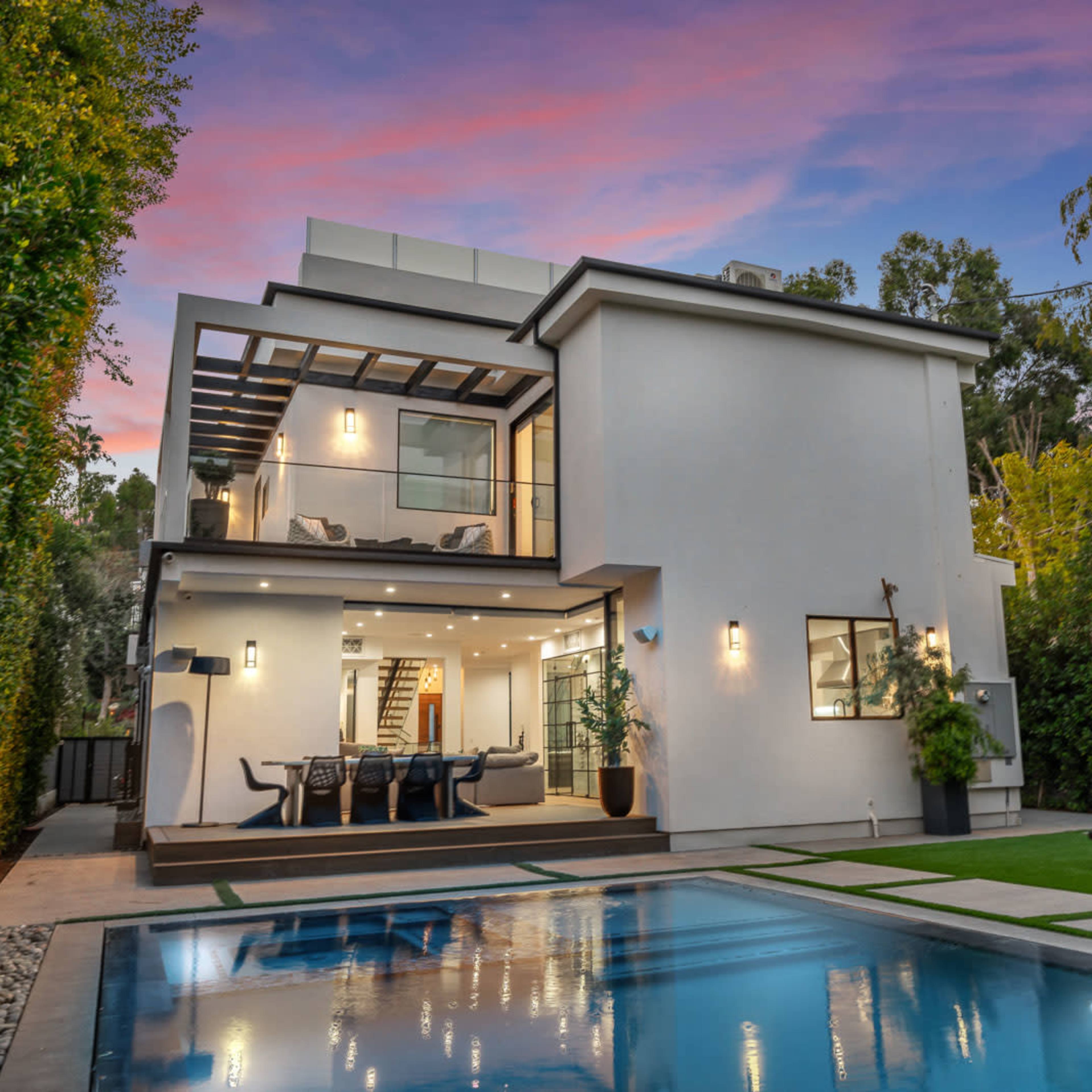 A modern two-story house with a swimming pool and outdoor seating area surrounded by greenery at sunset.