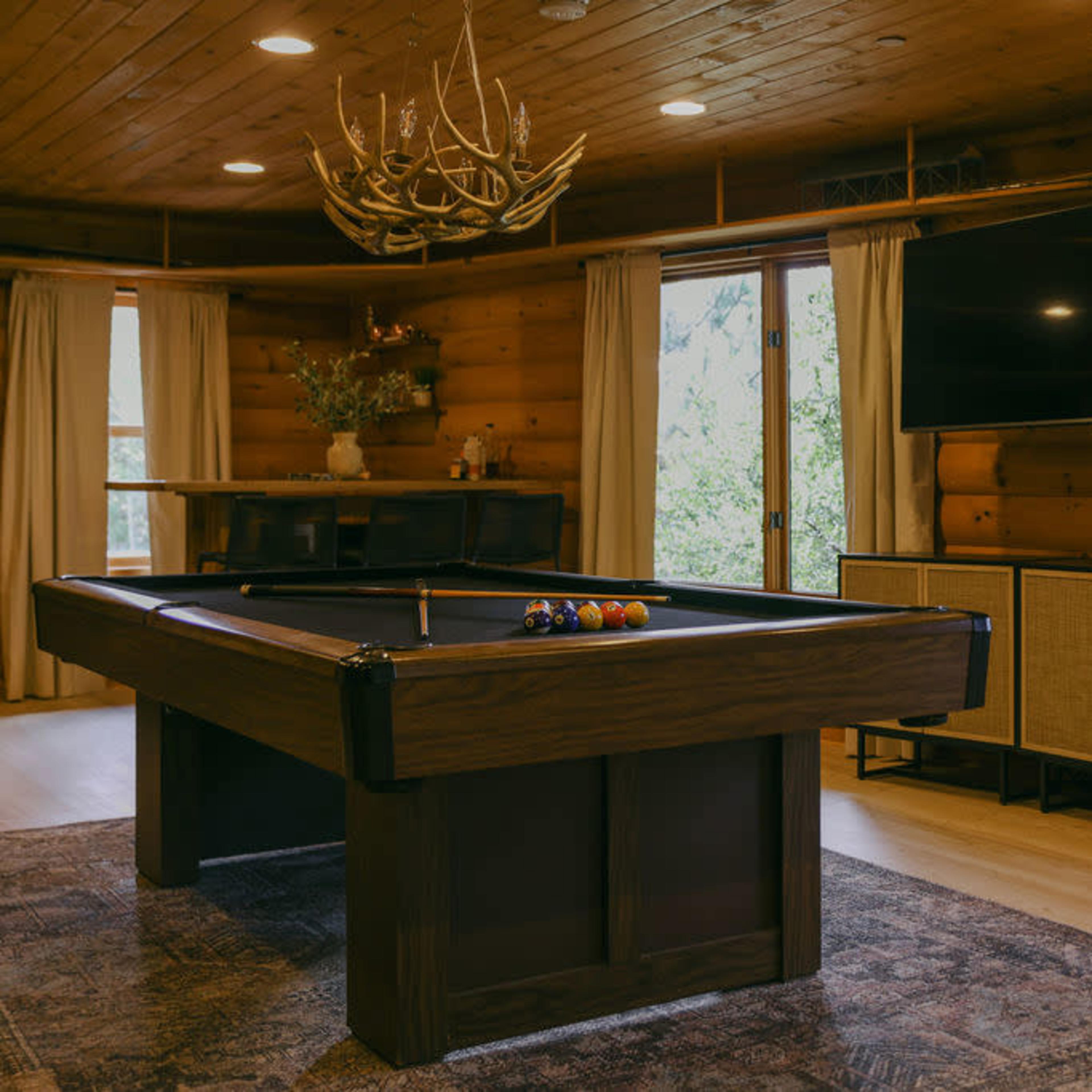 A rustic game room featuring a wooden pool table, a flat-screen TV mounted on the wall, and large windows with natural light illuminating the space.