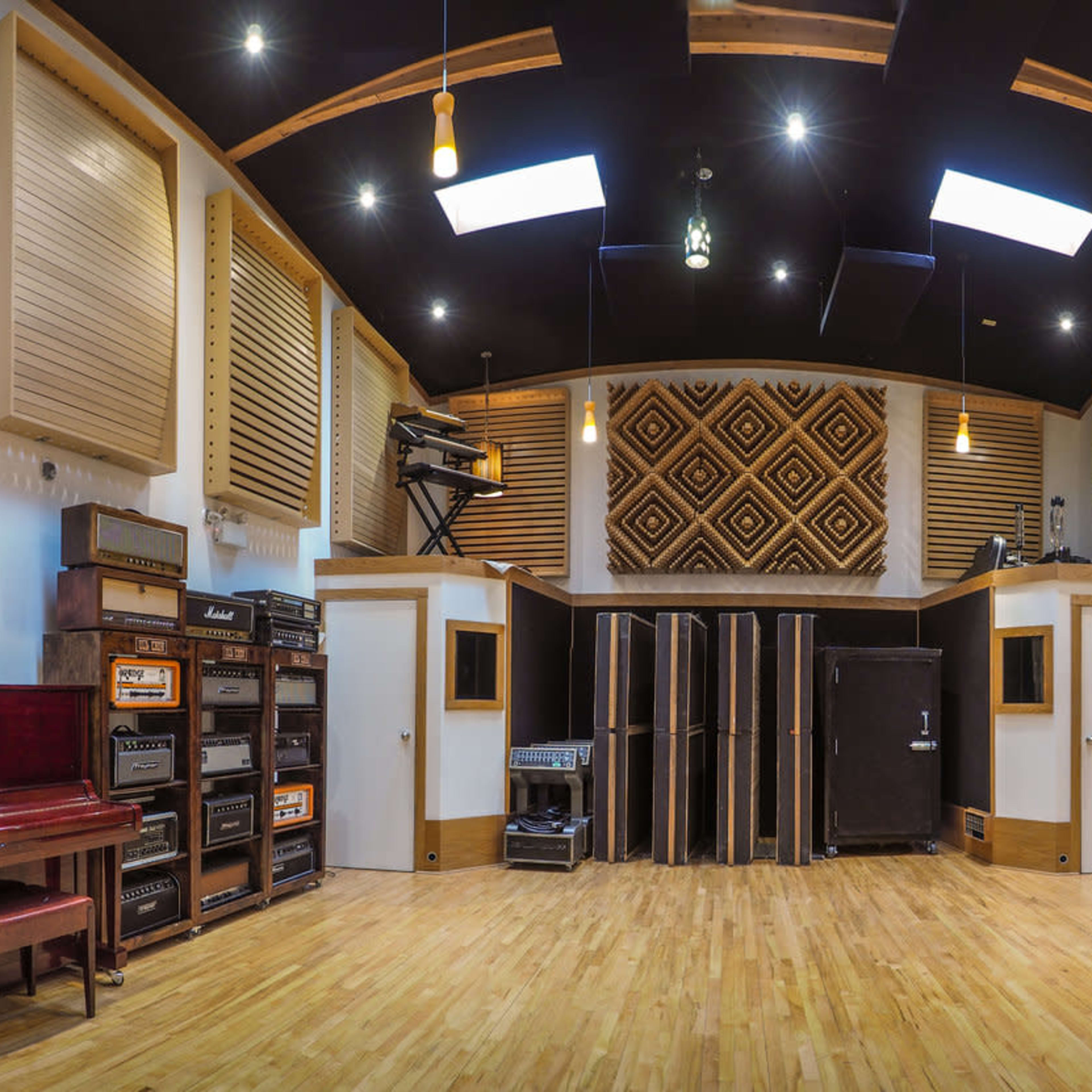 A spacious music studio with a wooden floor, soundproof walls, various amplifiers, a piano, and overhead lighting.