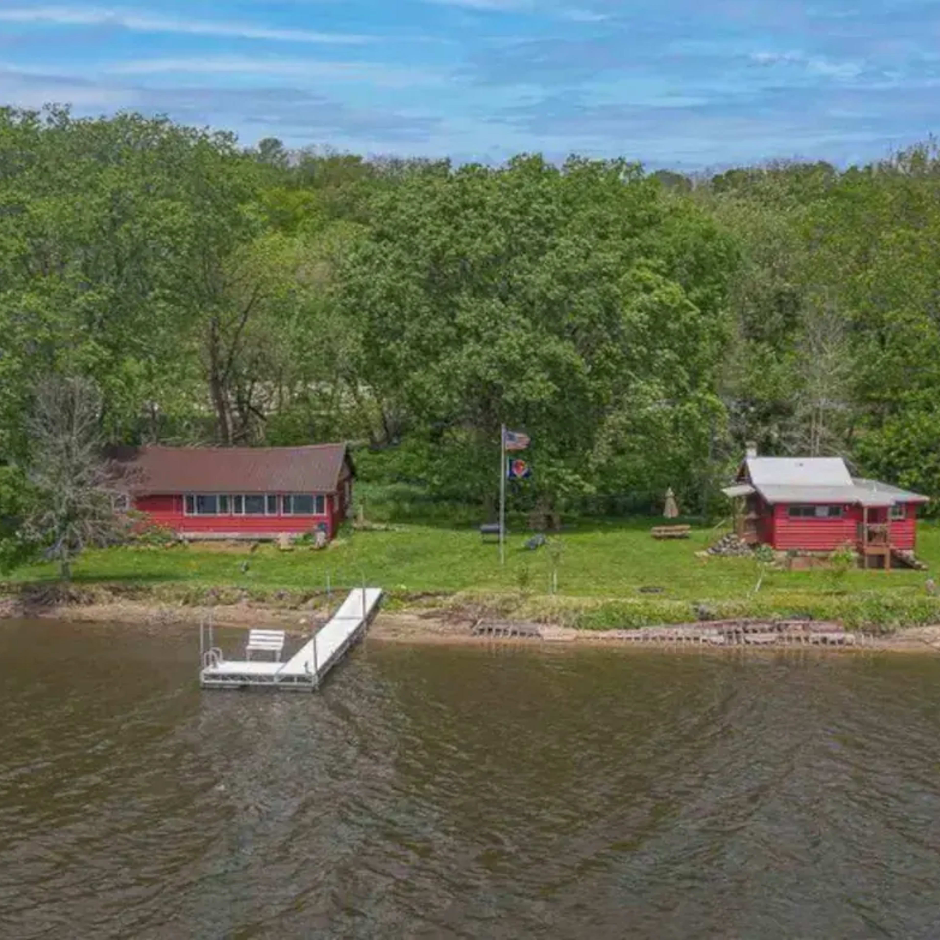 Two red cabins along a riverbank, surrounded by lush green trees, with a dock extending into the water.