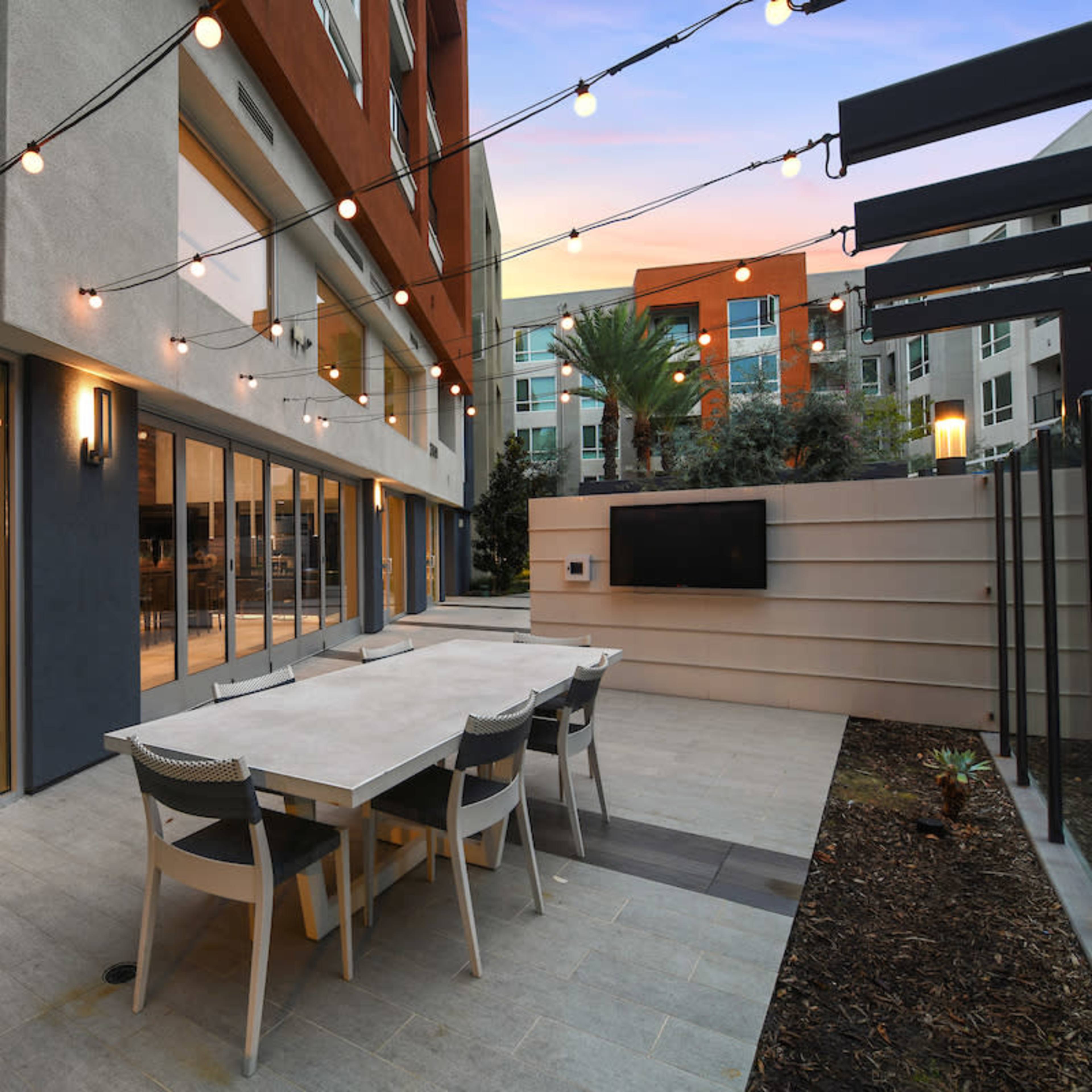 An outdoor patio area with a long dining table, surrounded by string lights and modern apartment buildings in the background.