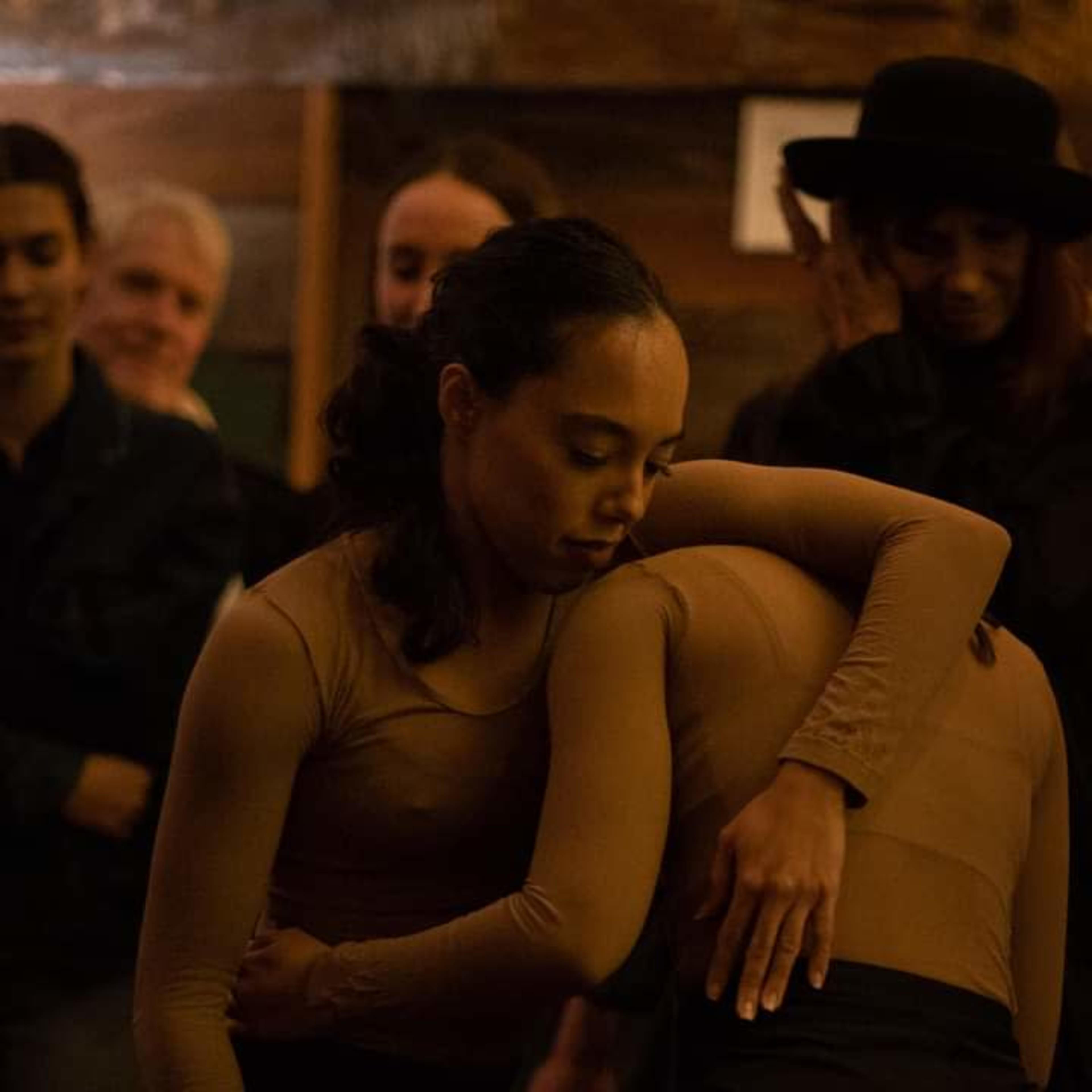 Two dancers embrace in a dimly lit room while others stand in the background.