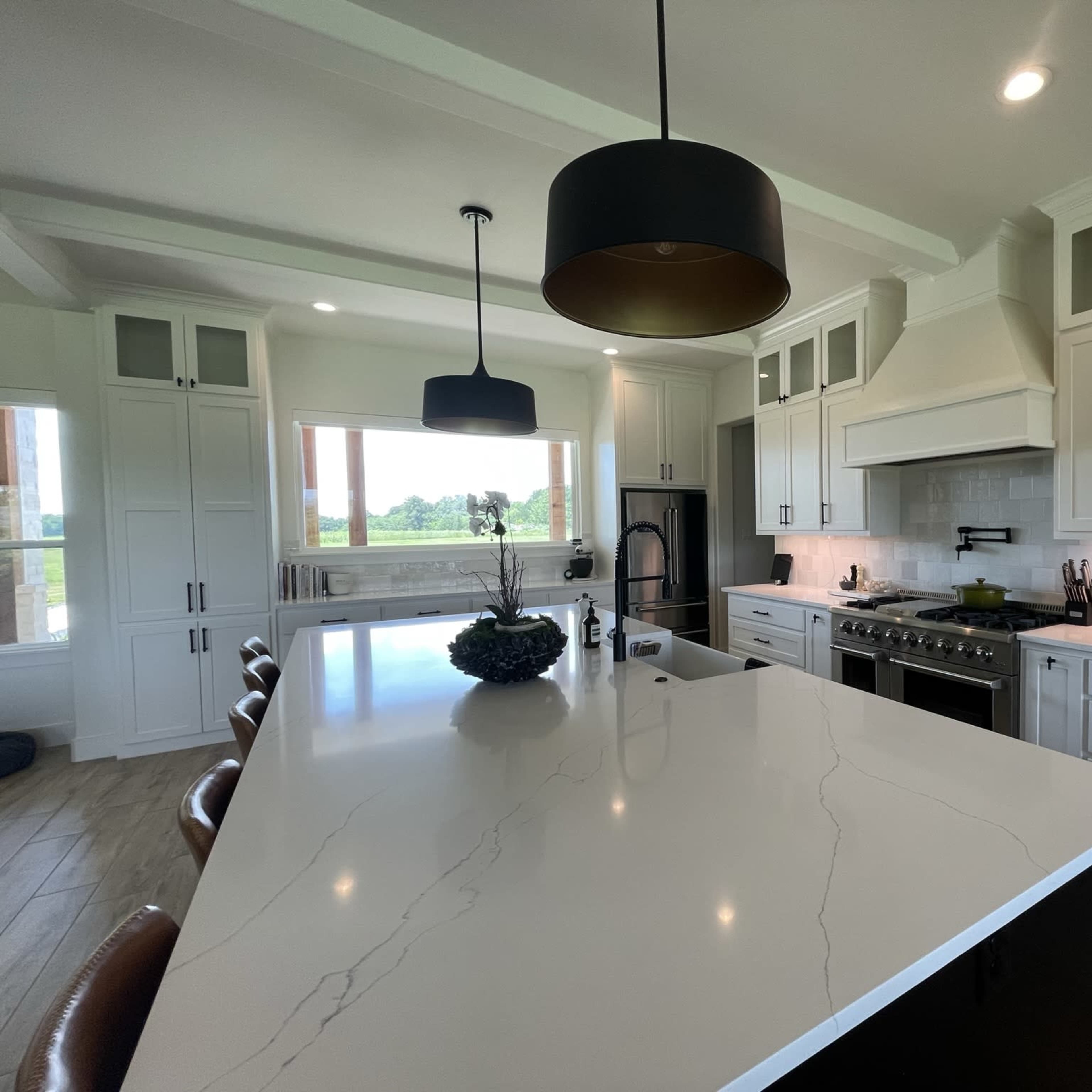A modern kitchen featuring a large island with a white countertop, dark cabinetry, and stainless steel appliances, set against a backdrop of large windows offering a view of the outdoors.