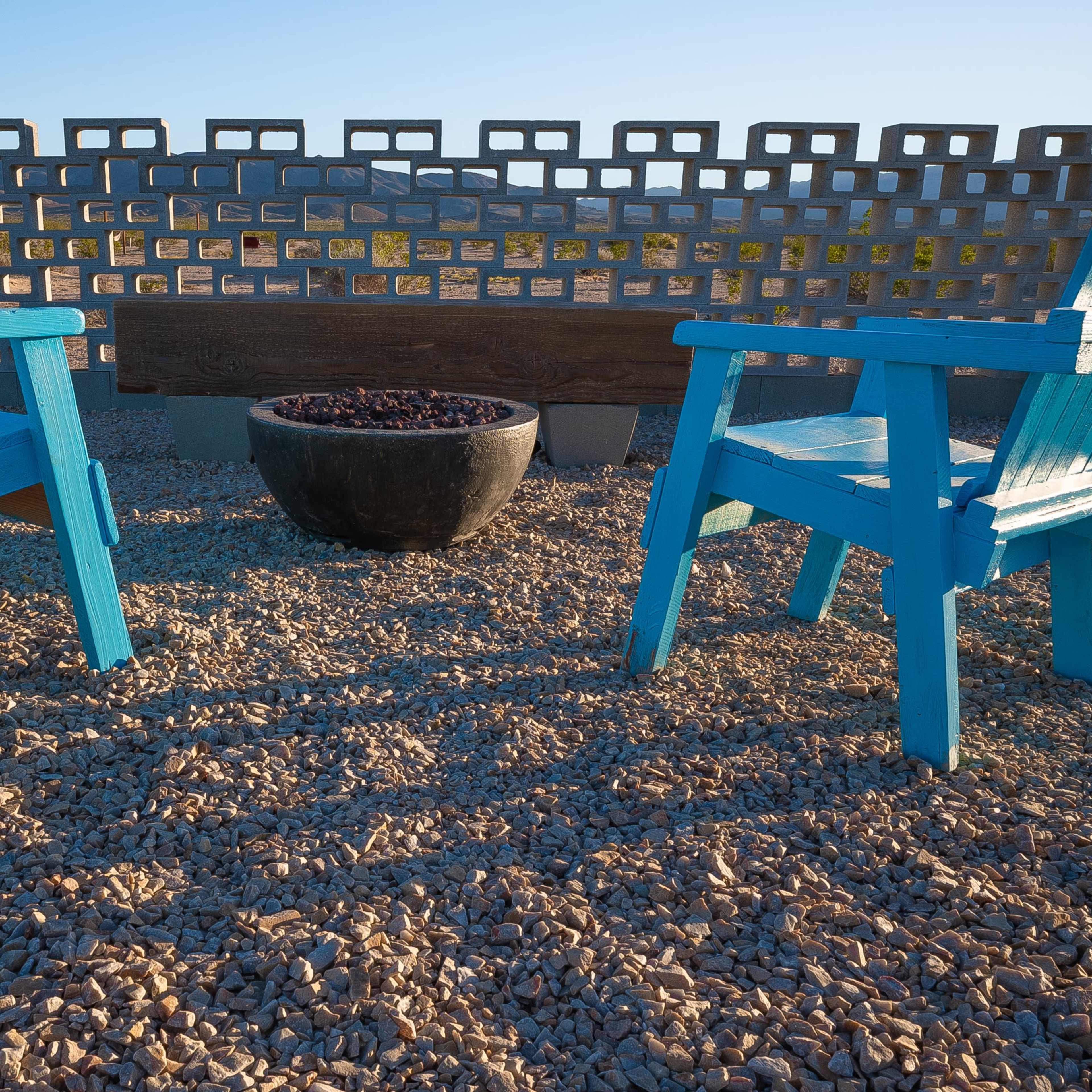 Two blue wooden chairs face a circular black planter, set on a gravel surface against a backdrop of a patterned concrete wall.