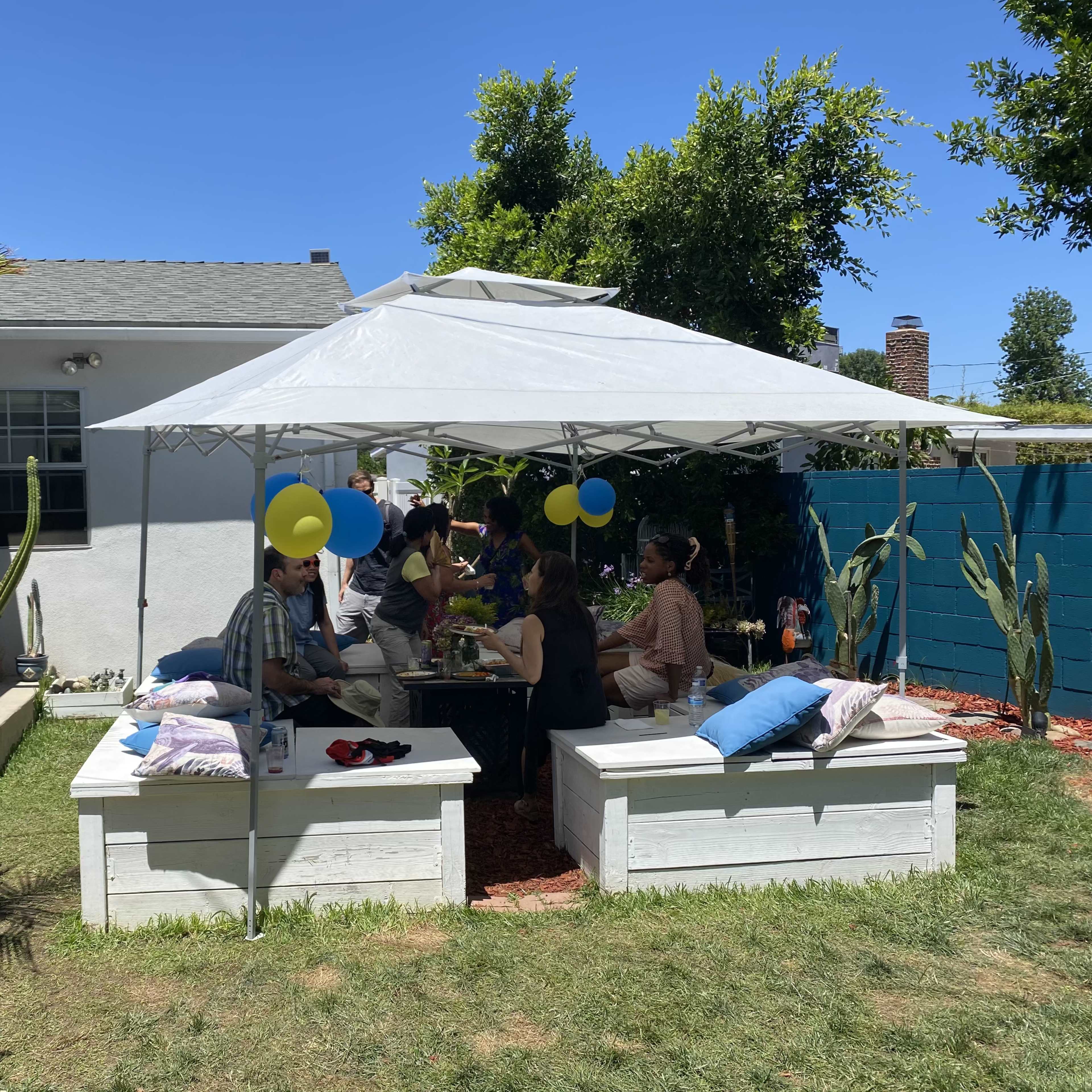 A group of people is dining under a white canopy in a backyard decorated with balloons and surrounded by green plants and a blue fence.