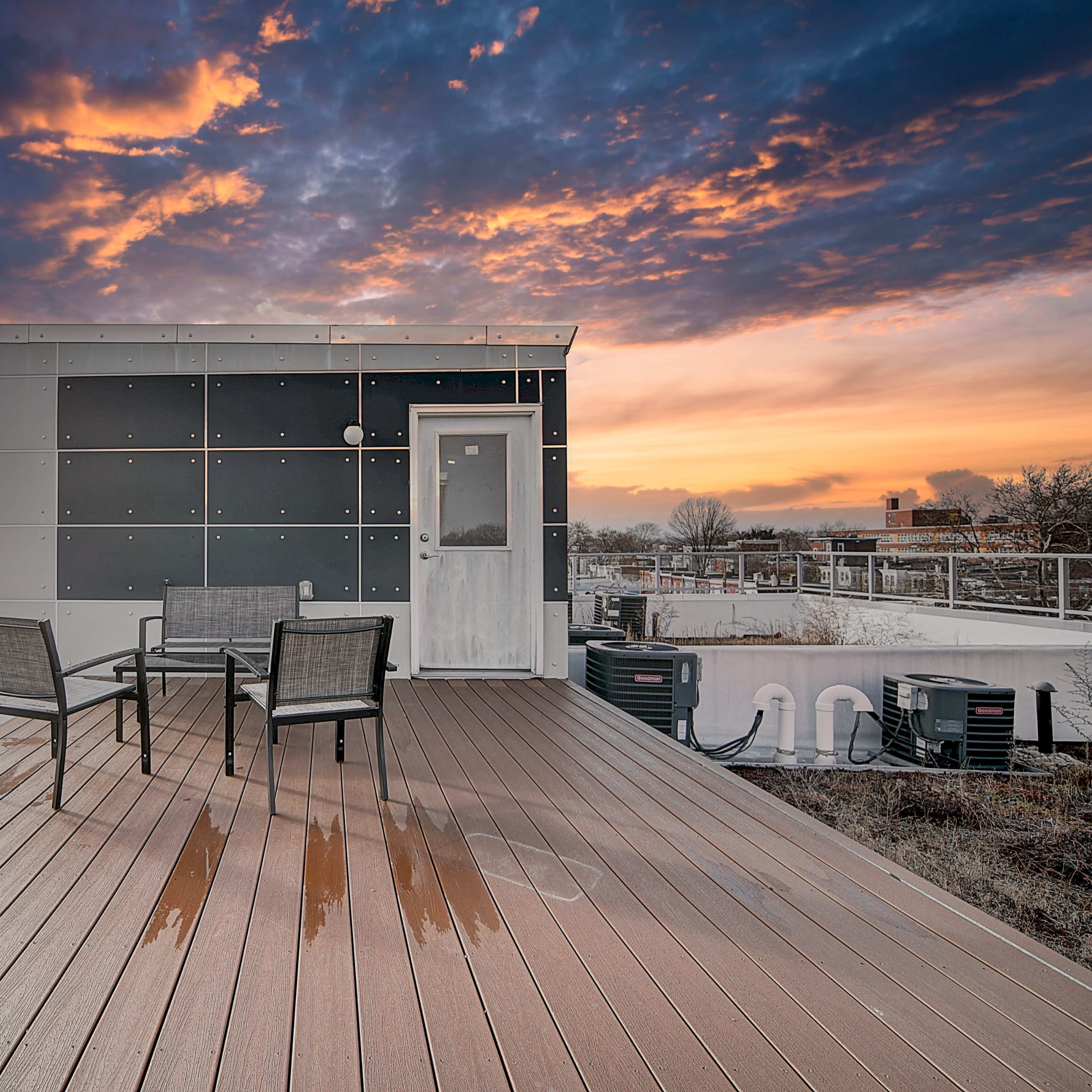 A rooftop deck features four chairs arranged around a small table, with a modern building and a colorful sunset in the background.