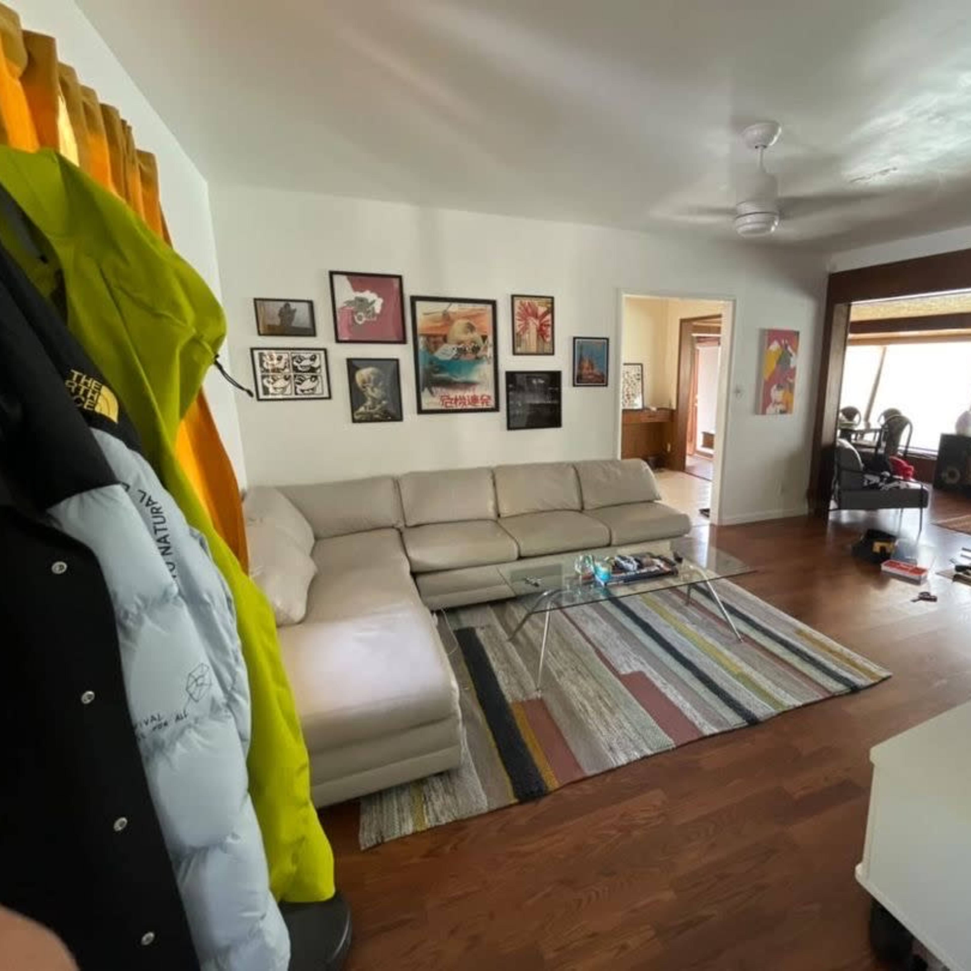 A living room with a sectional sofa, a glass coffee table on a striped rug, framed artwork on the walls, and various items scattered on the floor.