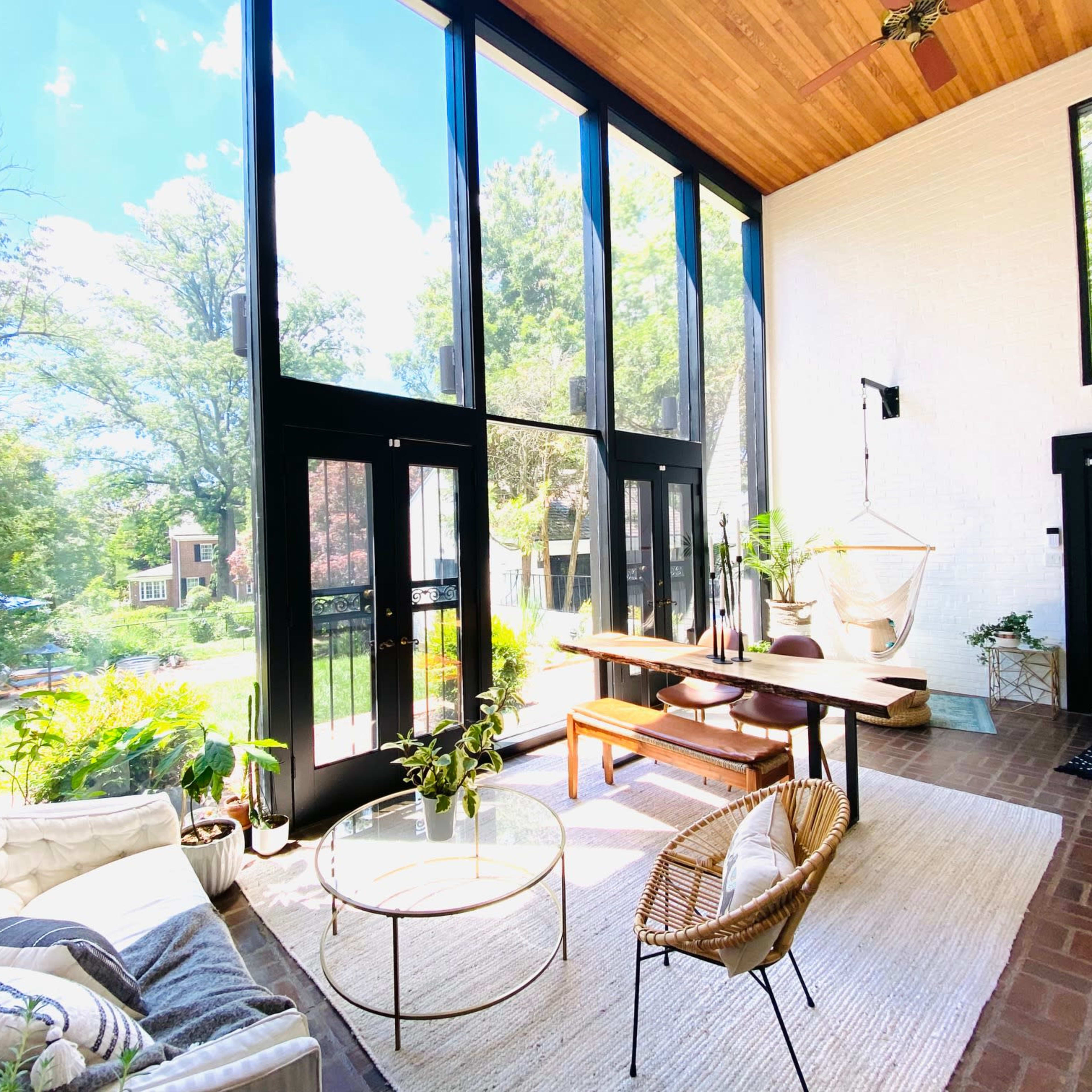 A bright and airy sunroom features large glass windows, a wooden ceiling, a dining table, and various plants.