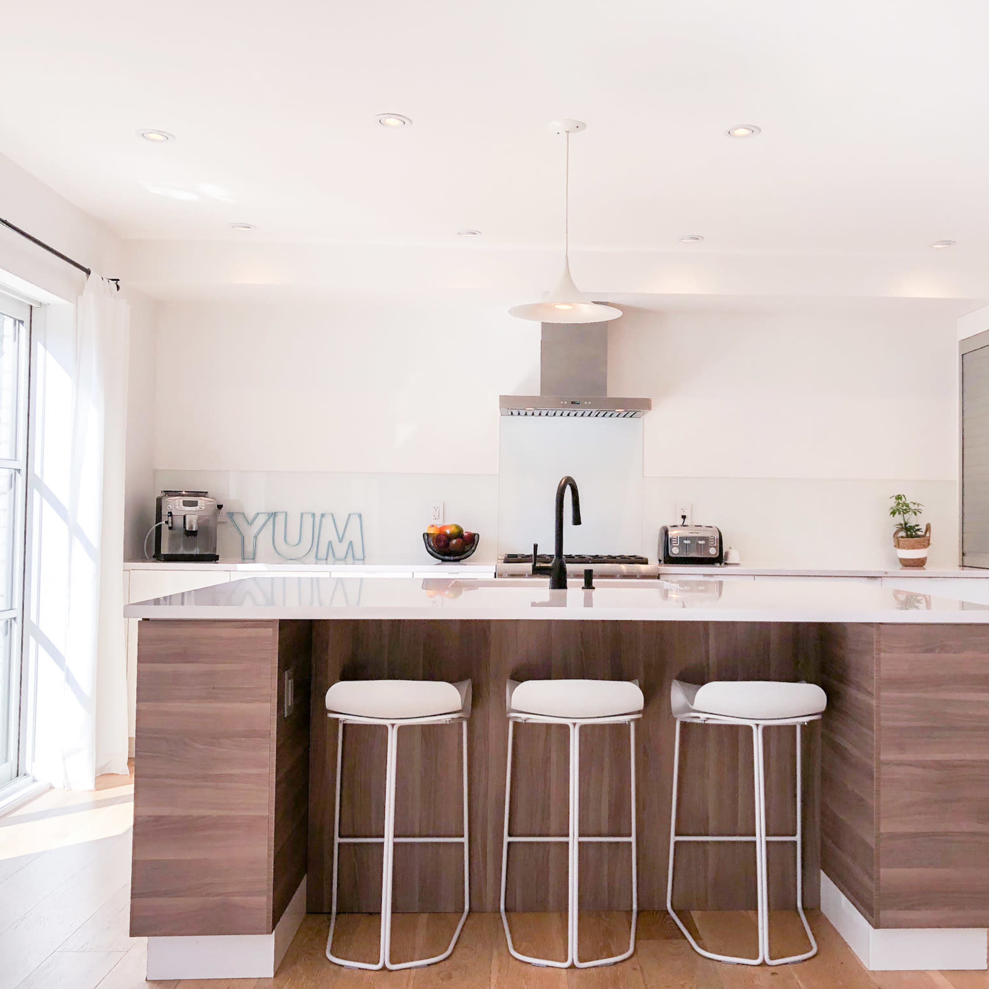 A modern kitchen features a central island with three bar stools, stainless steel appliances, and a sleek white countertop.