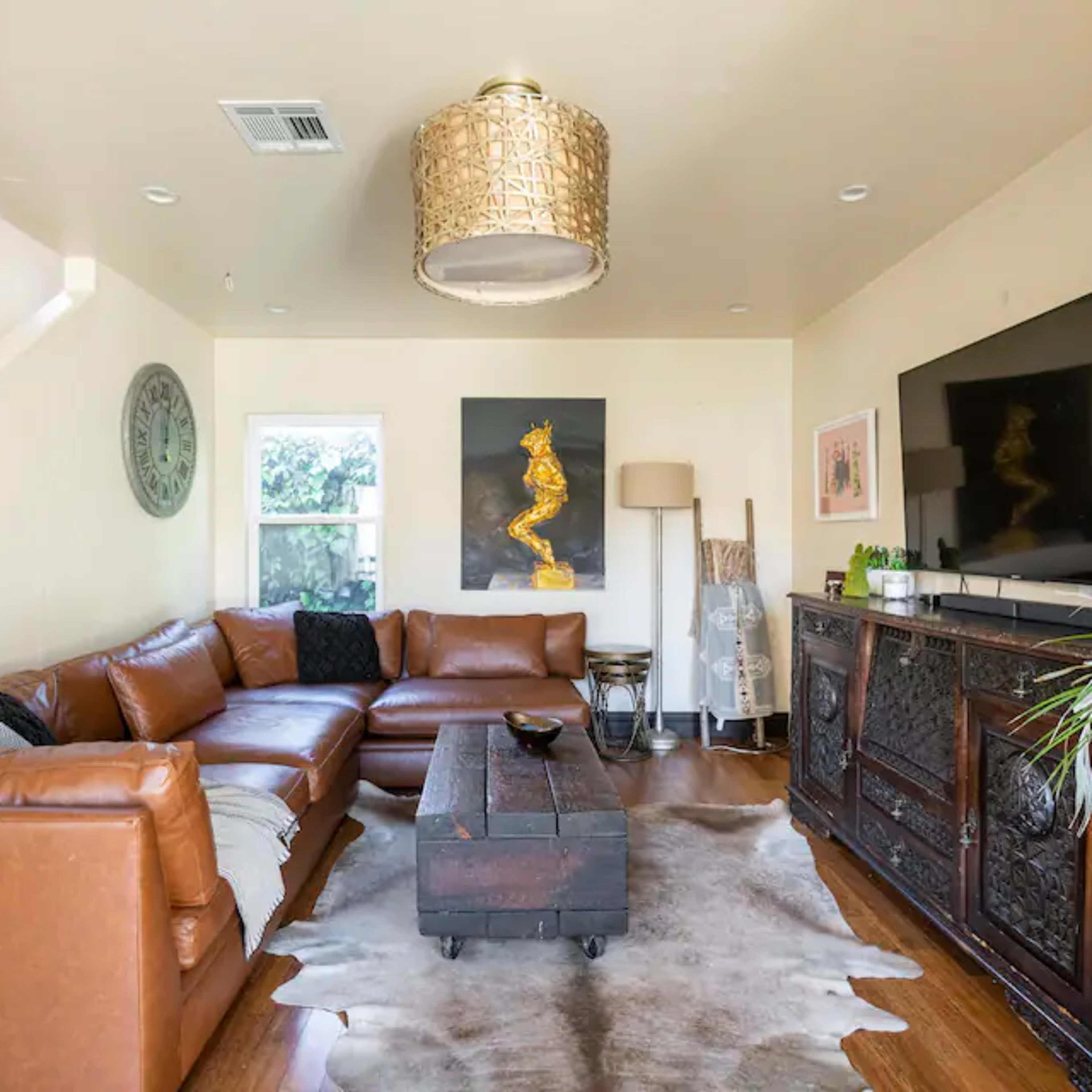 A spacious living room features a brown leather sectional sofa, a wooden coffee table, and a mounted television, with decorative artwork and a staircase in the background.