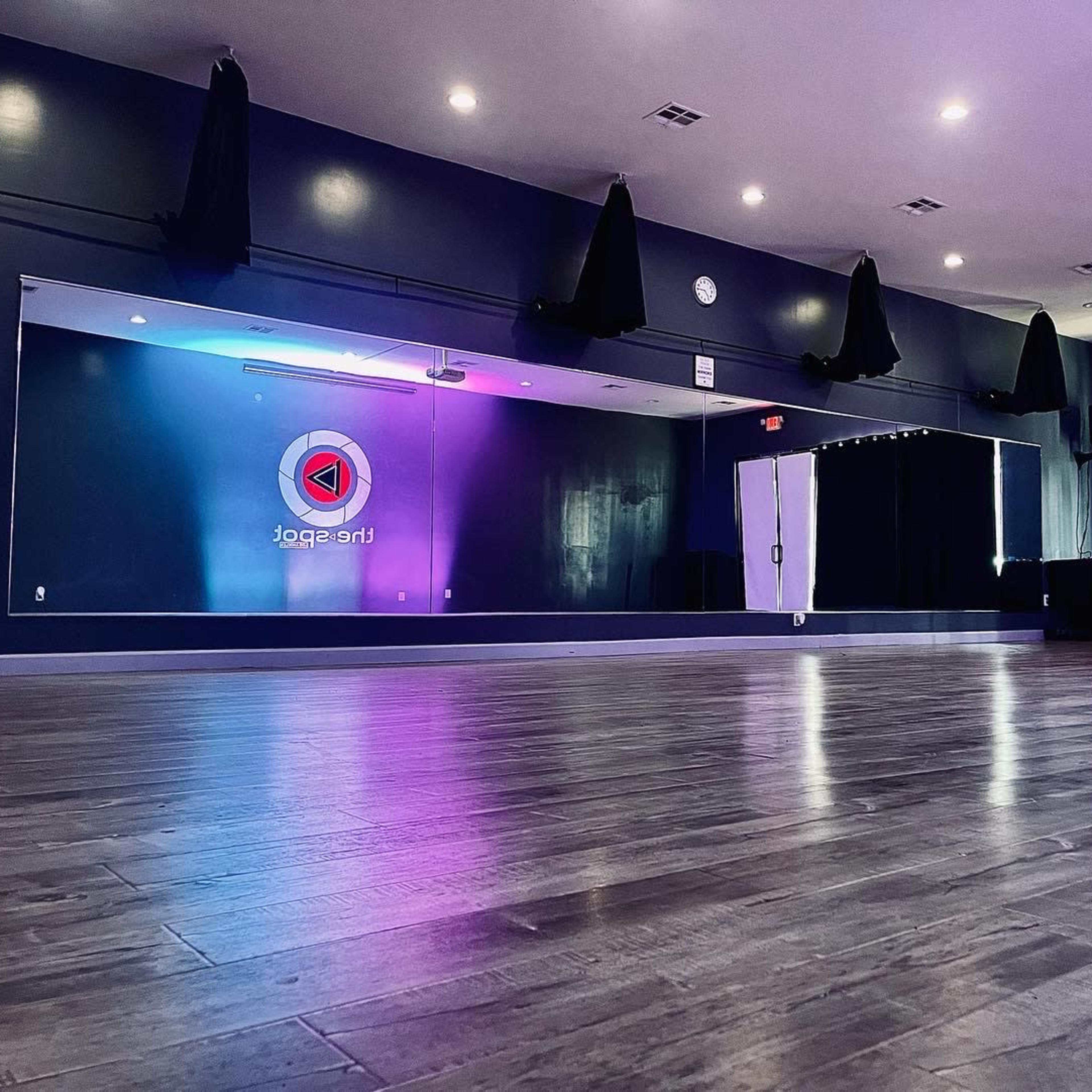 A spacious dance studio with a reflective wooden floor, large windows covered by dark curtains, and a colorful logo on the back wall.