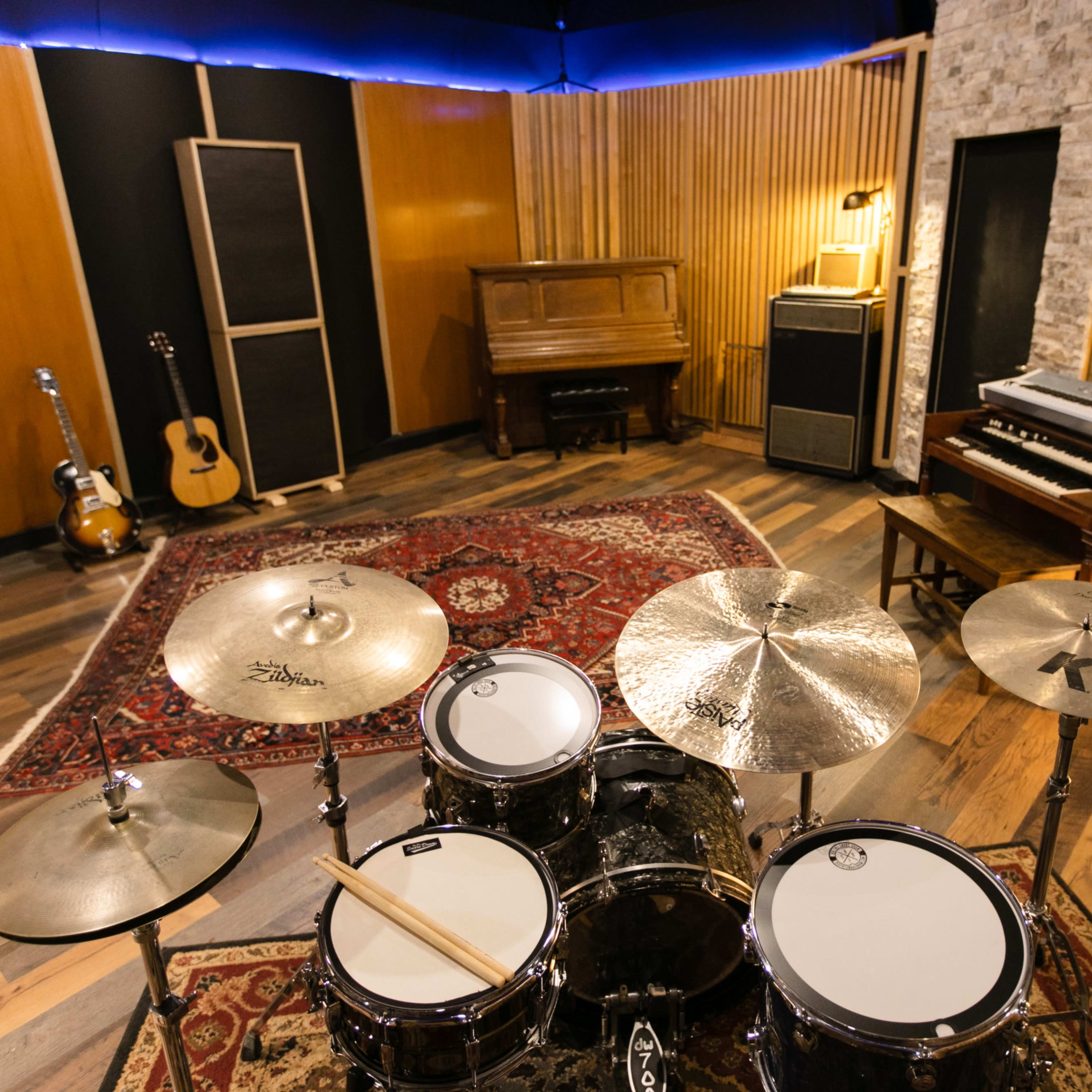 A music studio featuring a drum set in the foreground, a piano and a vintage amplifier in the background, and acoustic treatment on the walls.