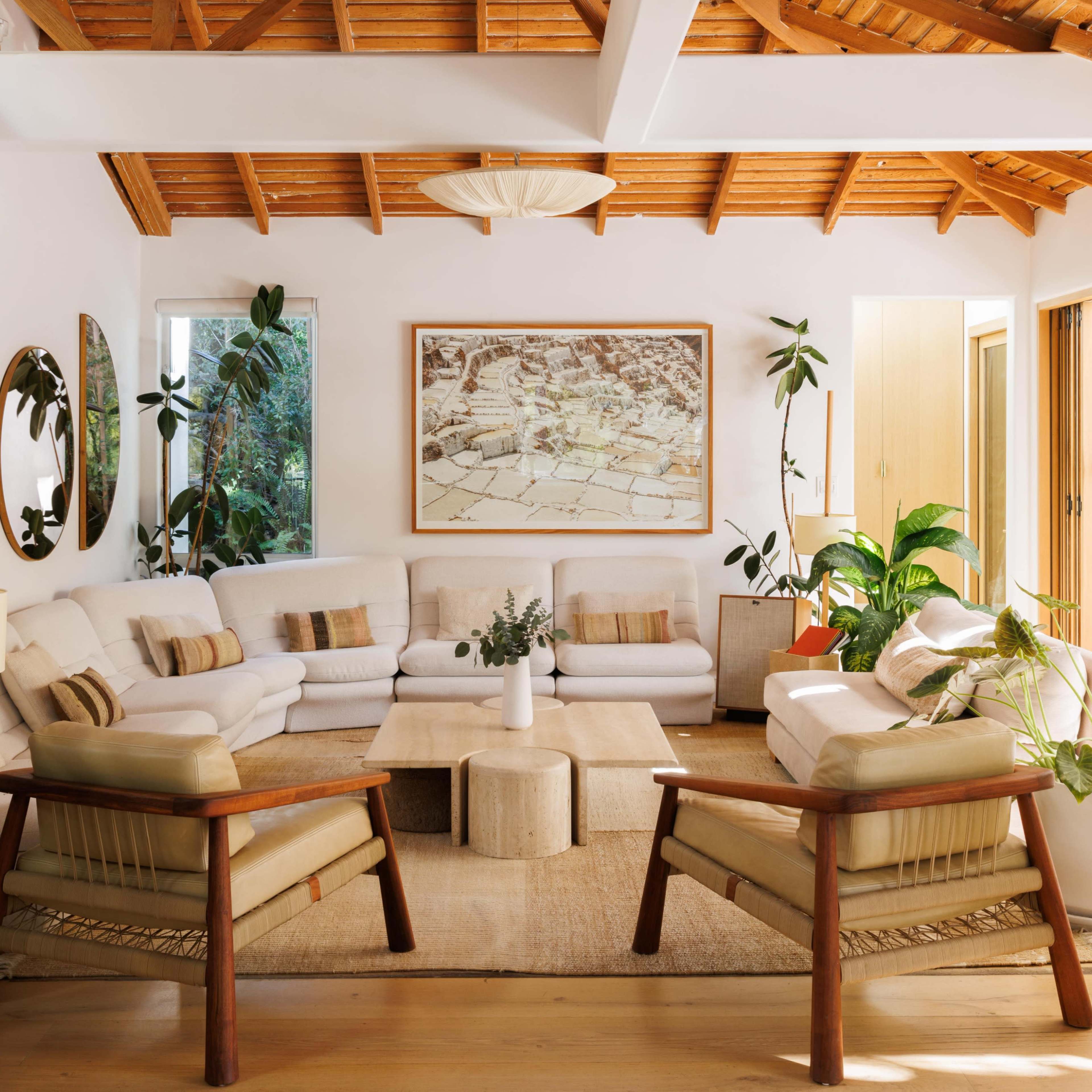 A modern living room with a high wooden ceiling, featuring white sofas, a light-colored coffee table, potted plants, and large windows opening to an outdoor area.