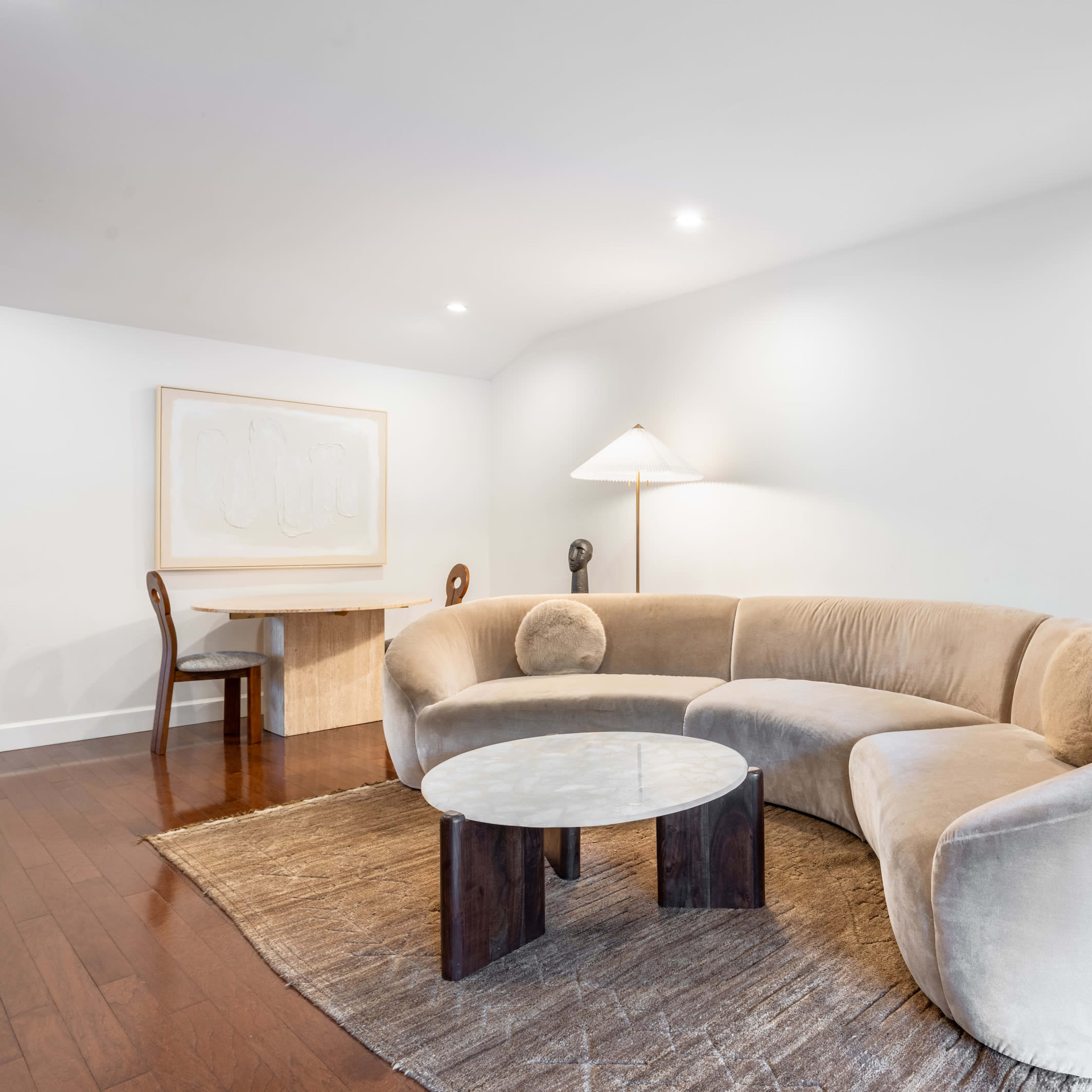 A modern living room featuring a curved beige sofa, a circular marble-top coffee table, a wooden desk, a lamp, and a potted plant in a decorative pot.