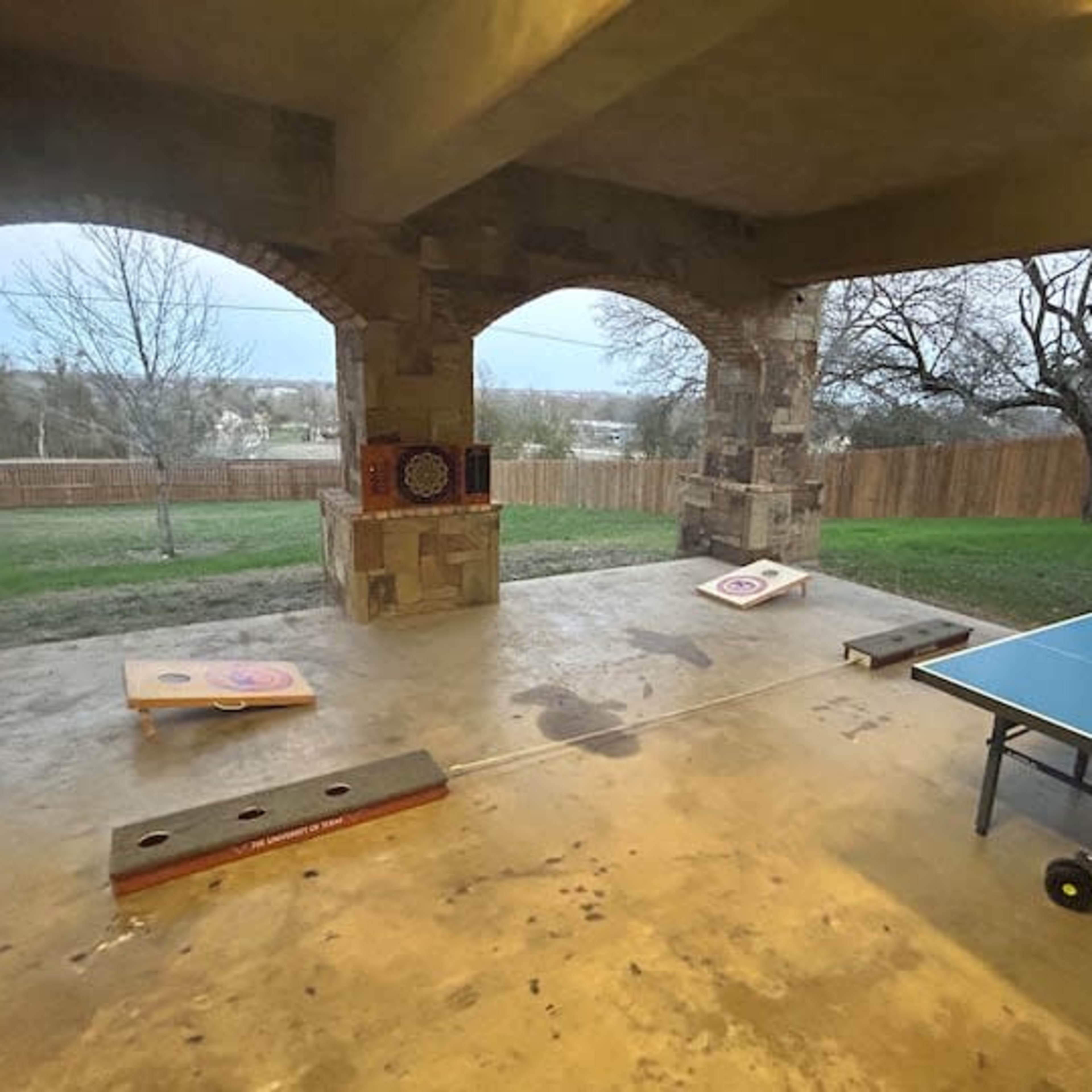 A covered outdoor area featuring a ping pong table and cornhole boards set up on a concrete floor, with a grassy landscape visible beyond the stone archways.