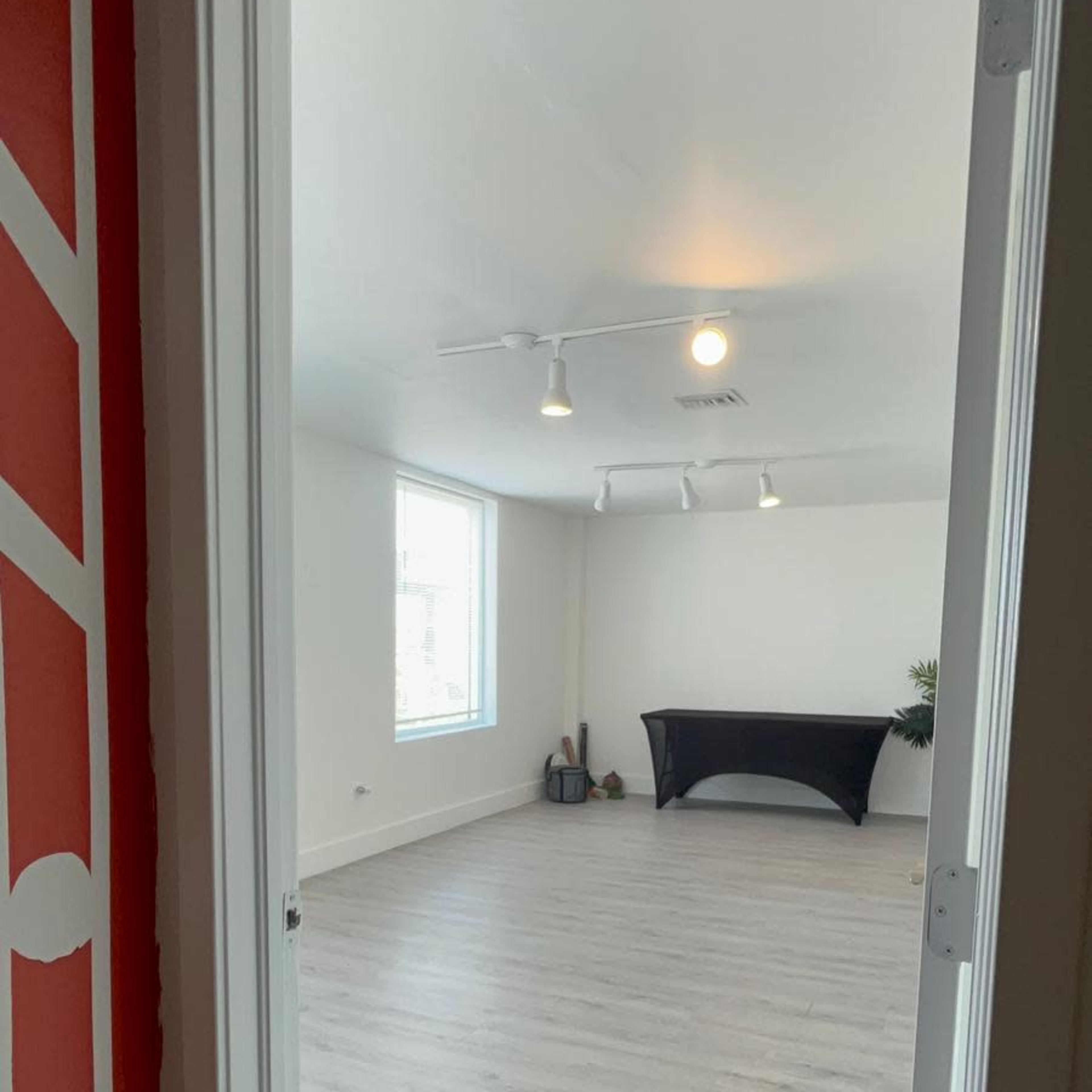 A doorway opens into a spacious room with light-colored flooring and a black table against one wall.