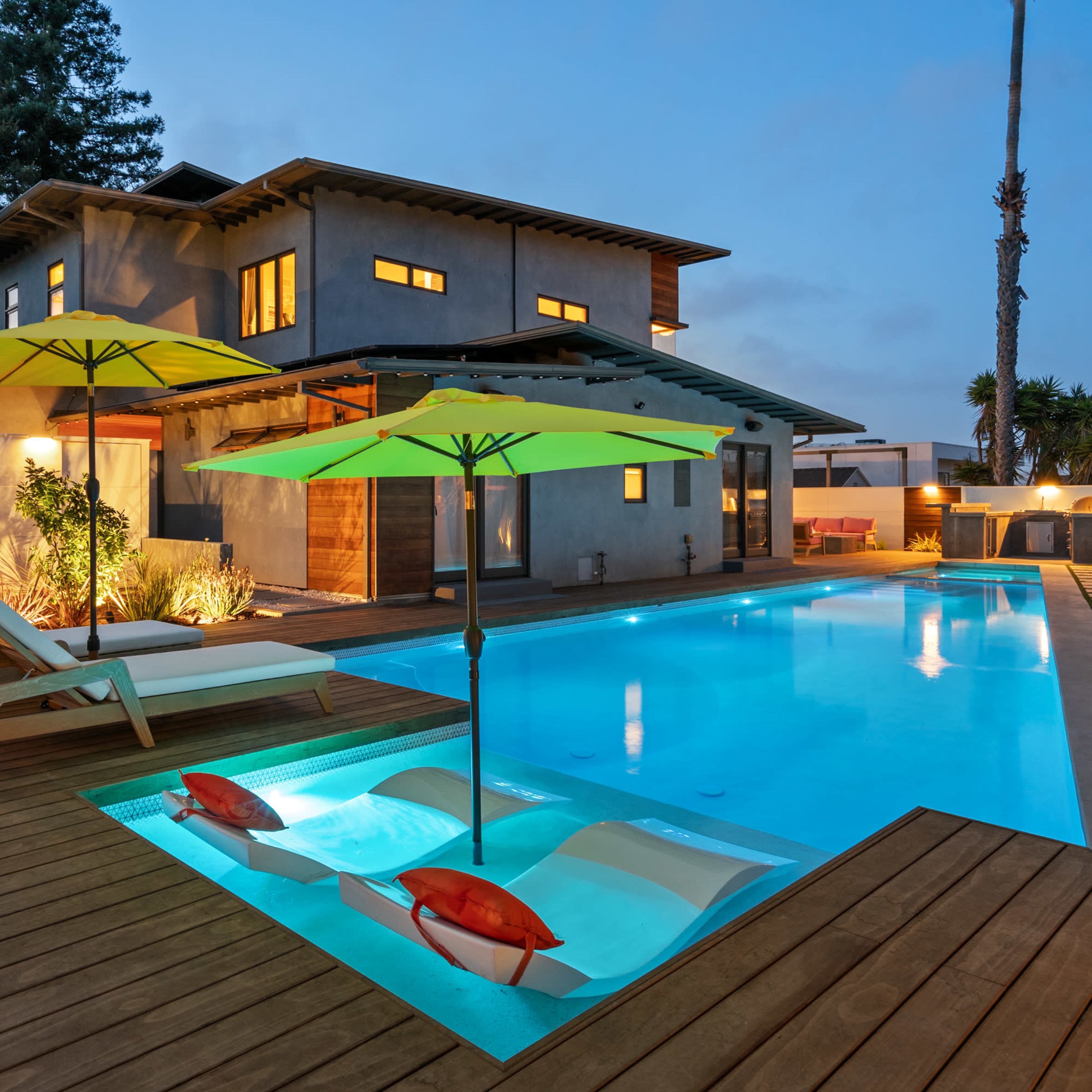 The scene depicts a modern home with a pool surrounded by wooden decking, featuring two lounge chairs and bright umbrellas.