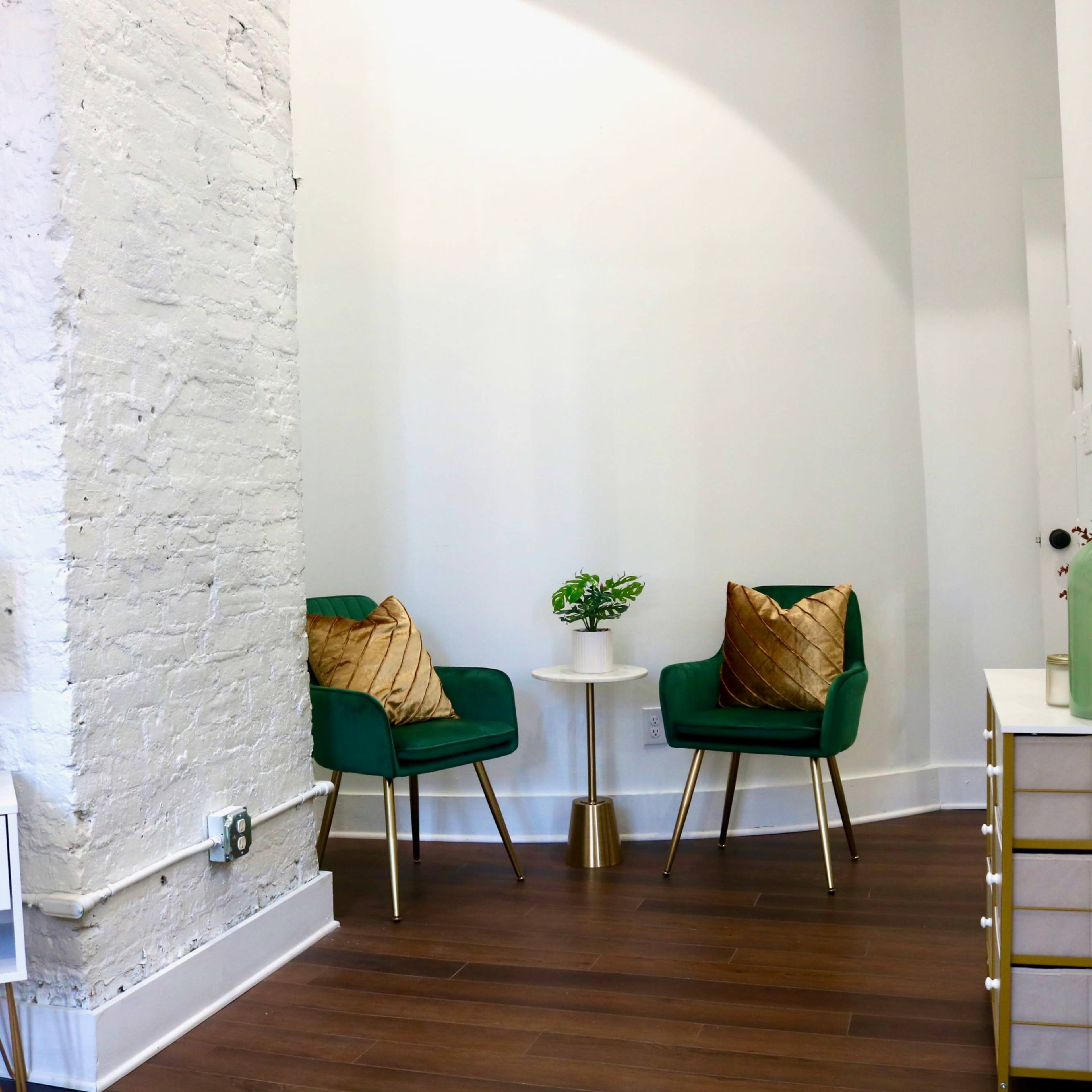 A corner of a modern room features two green chairs with gold accents, a small table between them, and a decorative mirror on the wall.