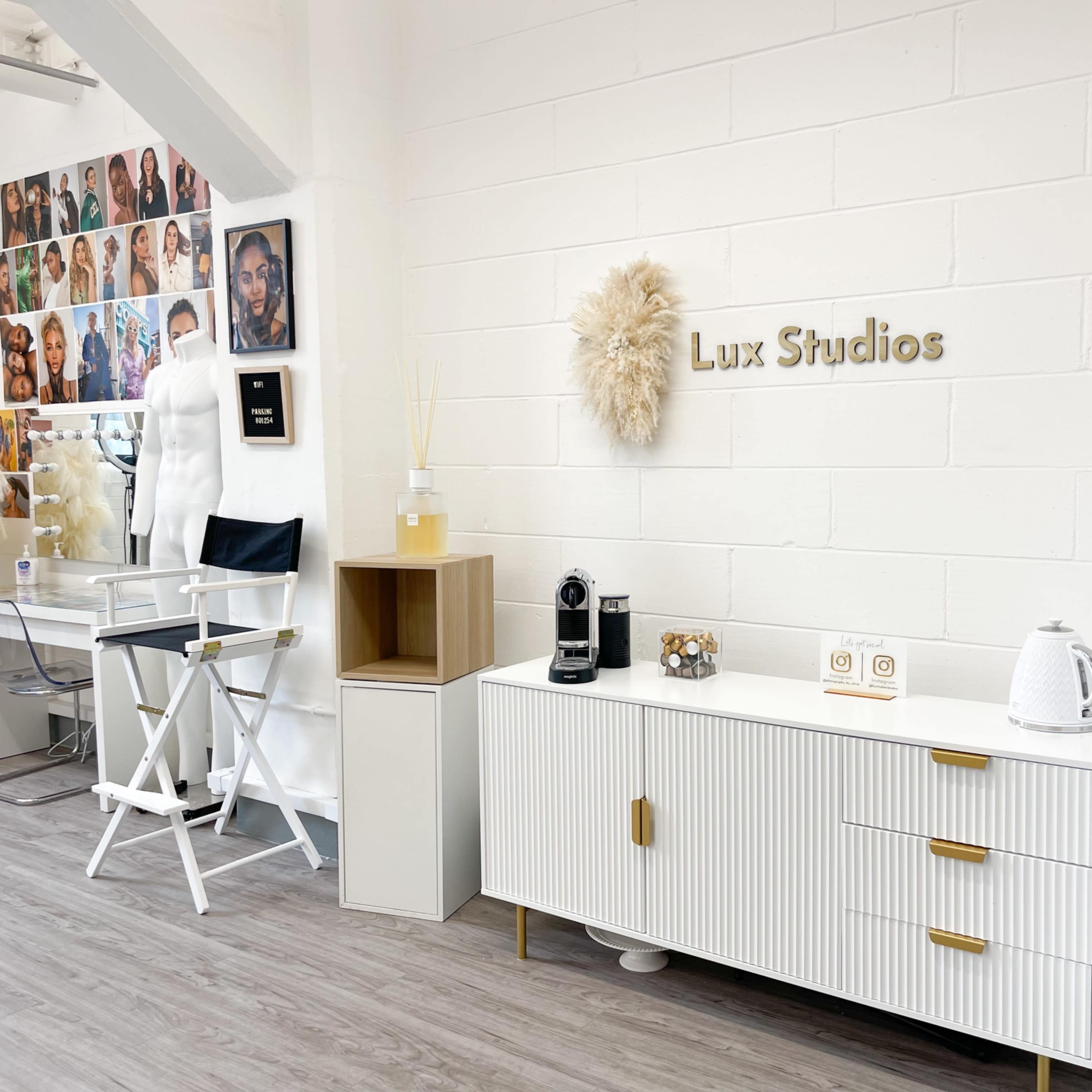A modern studio interior featuring a white wall with a decorative sign reading "Lux Studios," along with a cabinet, a coffee machine, and a gallery of portraits on the adjacent wall.