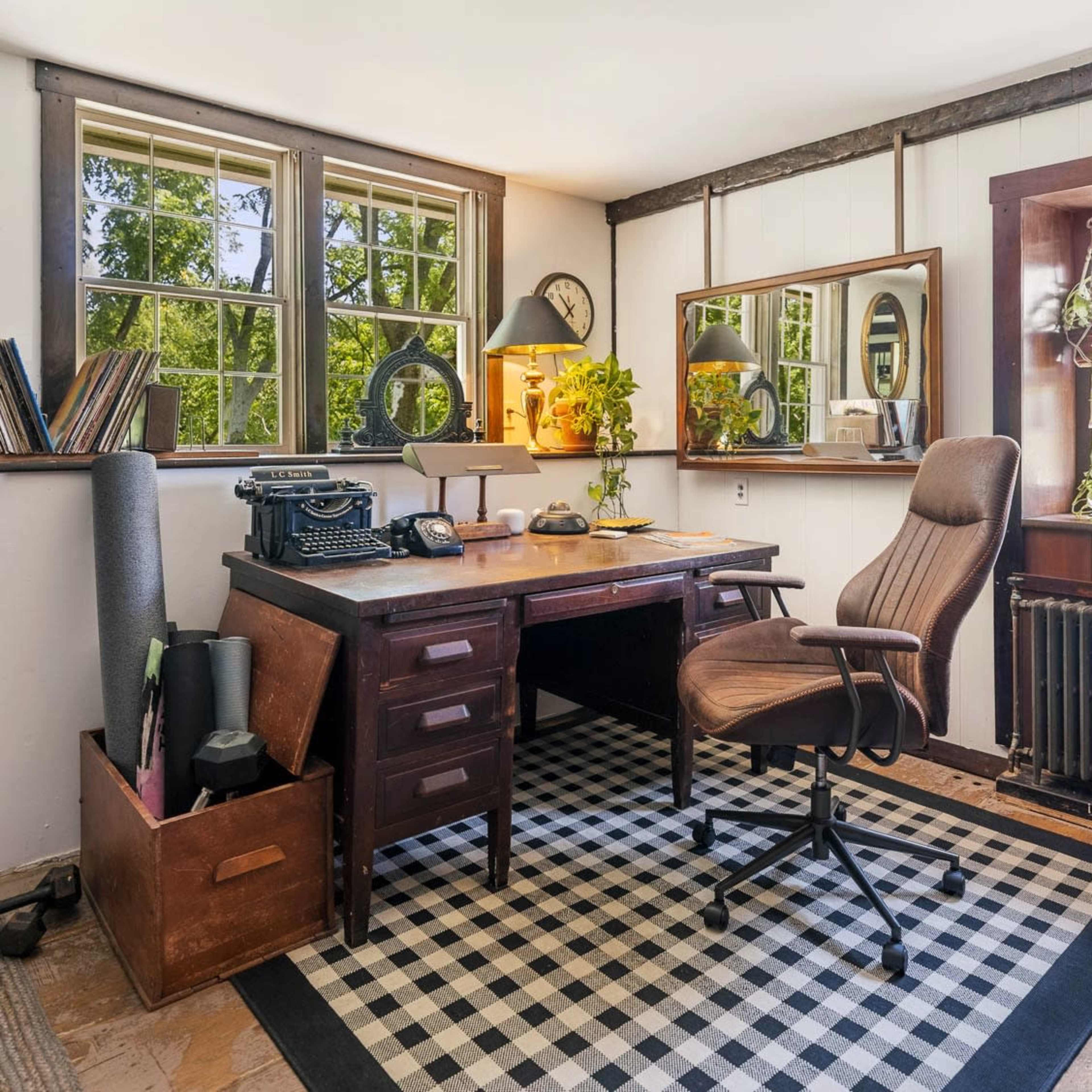 A vintage wooden desk with a typewriter and a chair is positioned in a sunlit room featuring large windows and decorative mirrors.