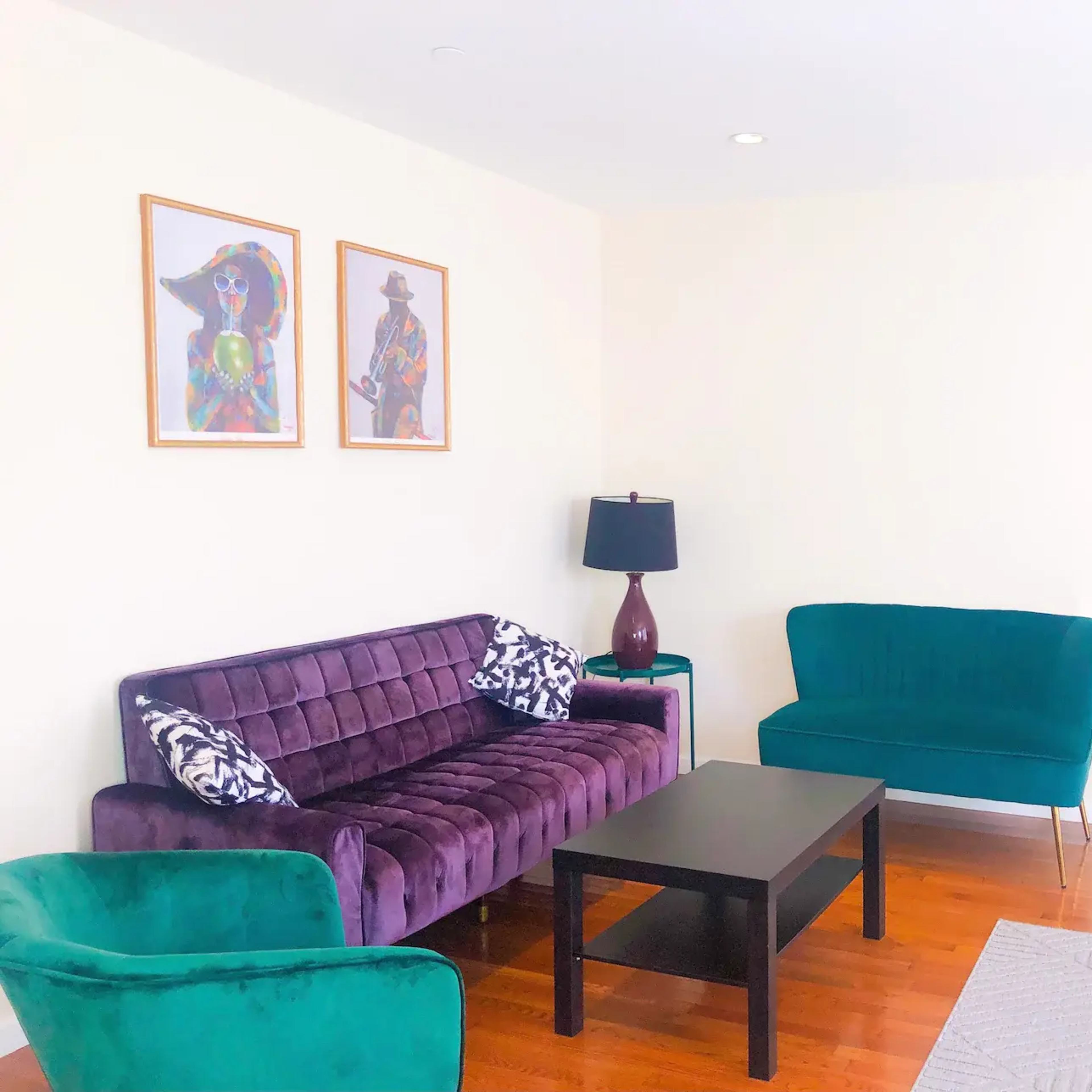 A living room featuring a purple velvet sofa, a green upholstered armchair, and a green loveseat, along with a black coffee table and wall art above the sofa.