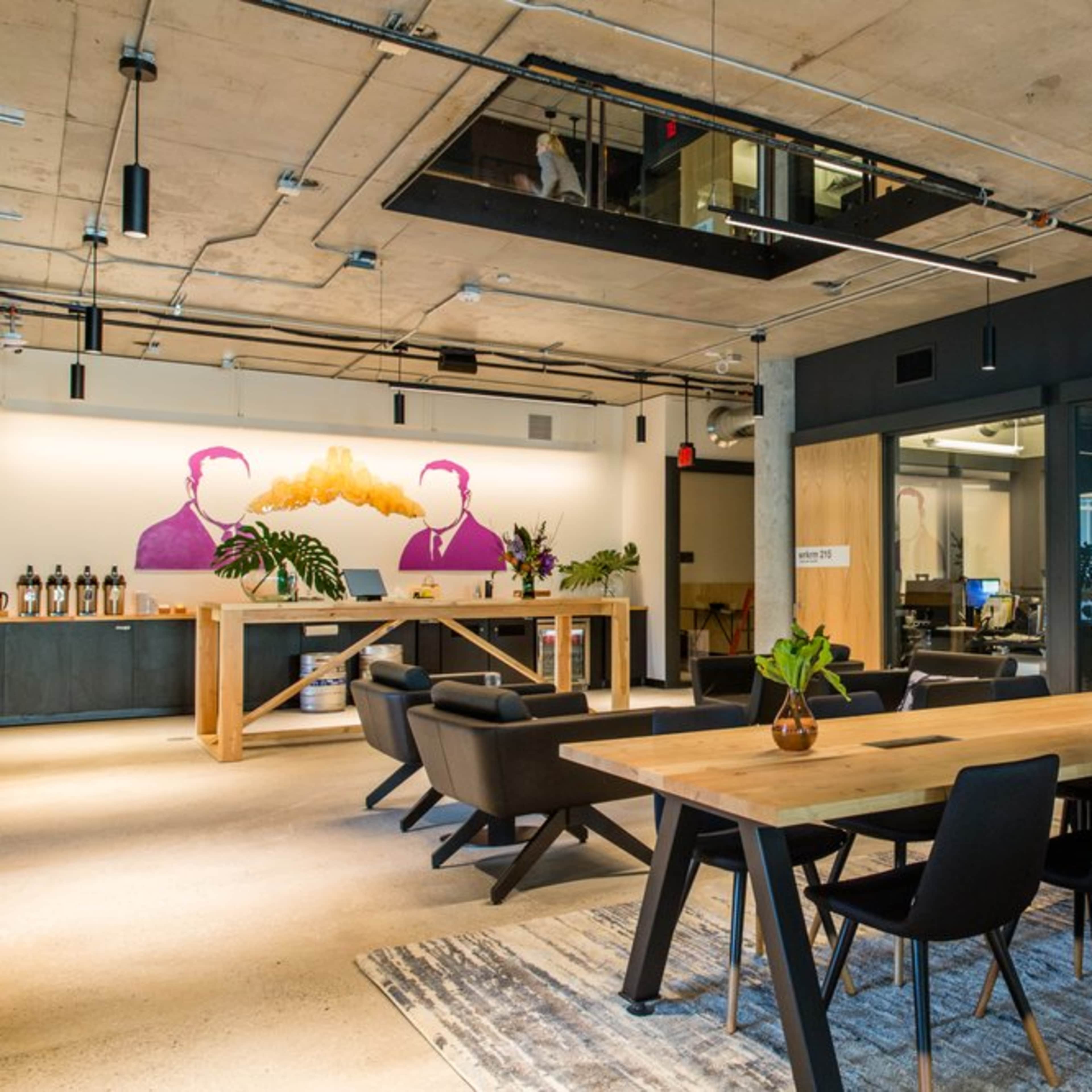 A modern office space with a large wooden table, black chairs, and a mural of two figures on the wall, alongside glass office partitions and greenery.