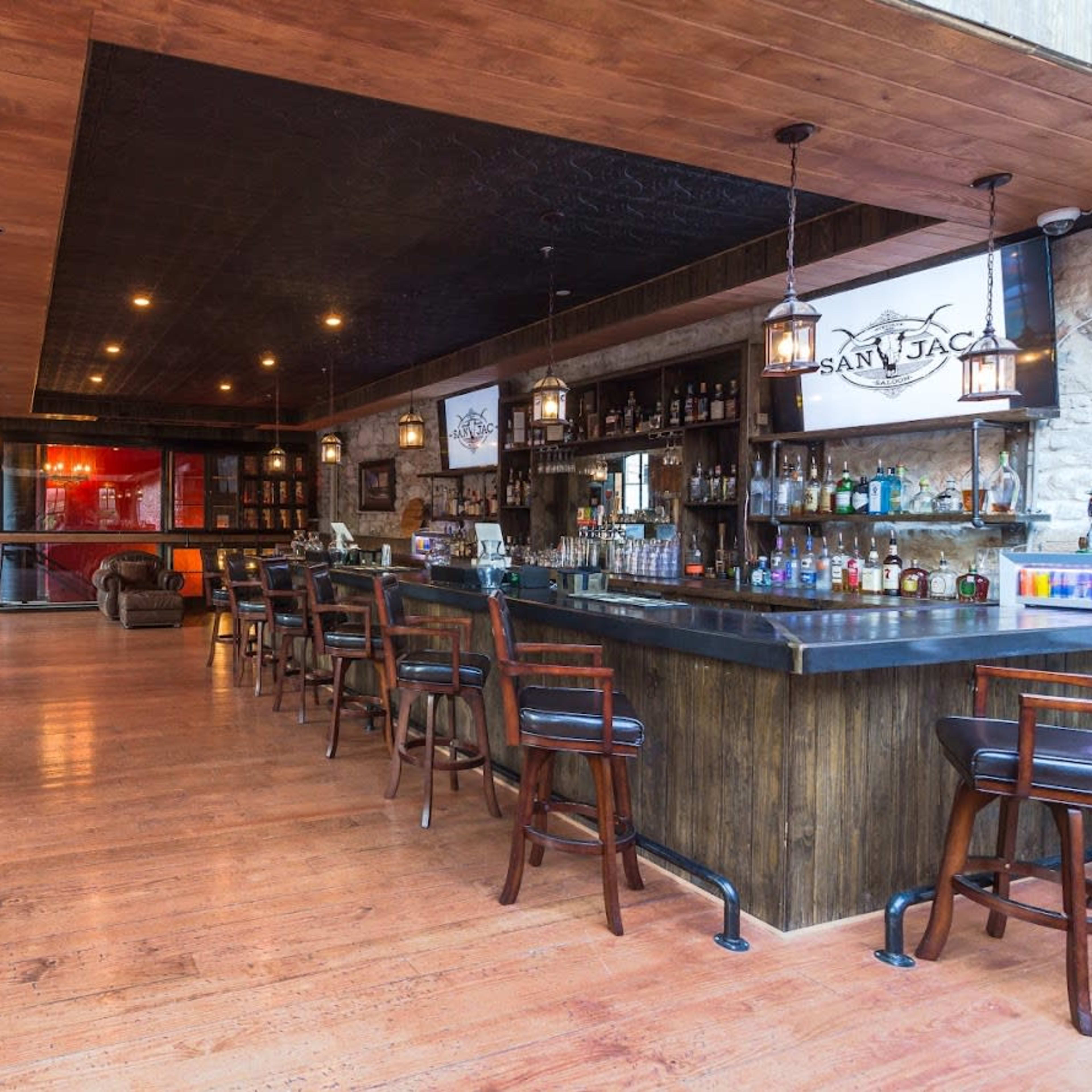 A bar interior with wooden floors, a large bar counter, and seating arrangements of tables and high chairs.