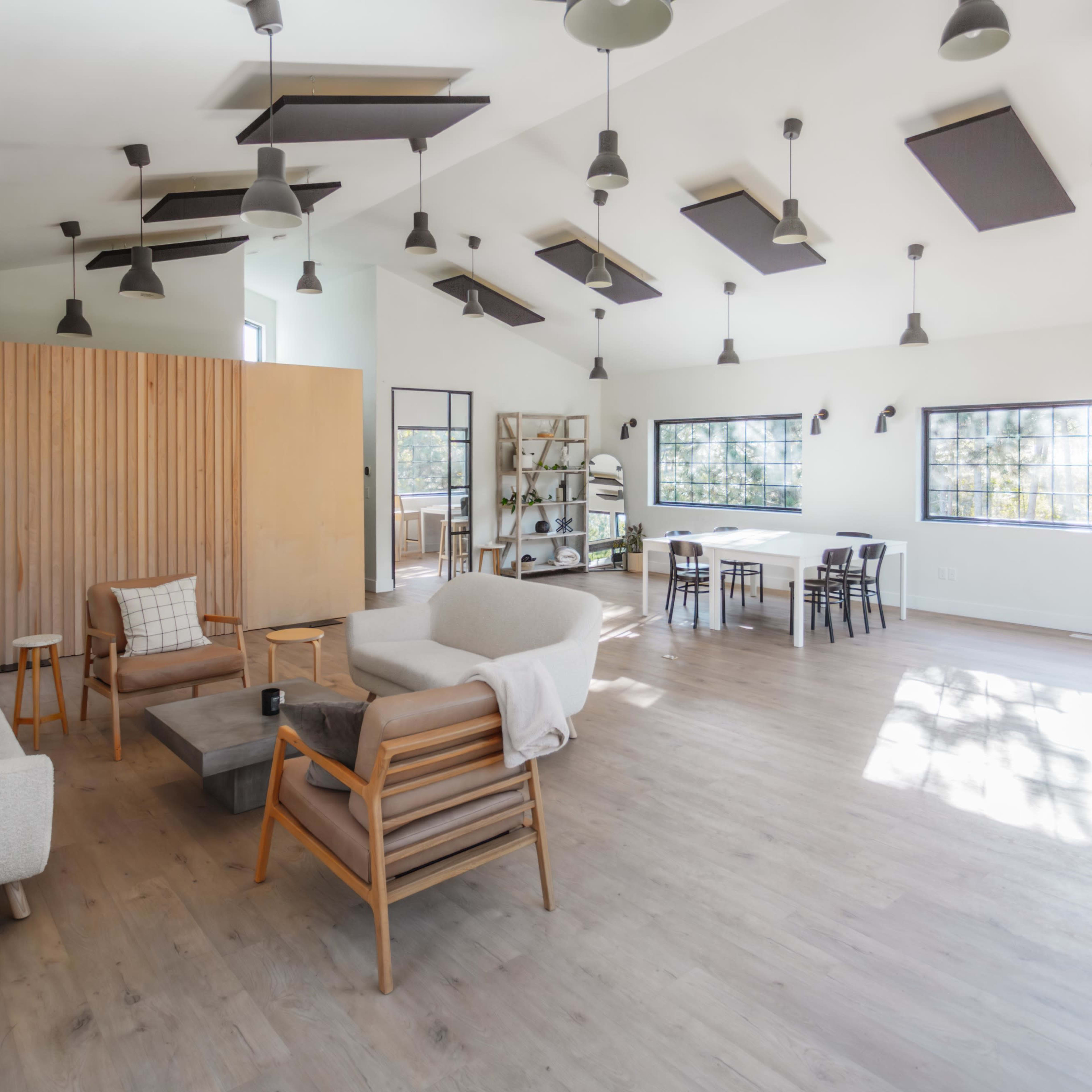 A spacious, modern interior of a bright room with wooden flooring, featuring a seating area with sofas, a wooden partition, and a dining table.