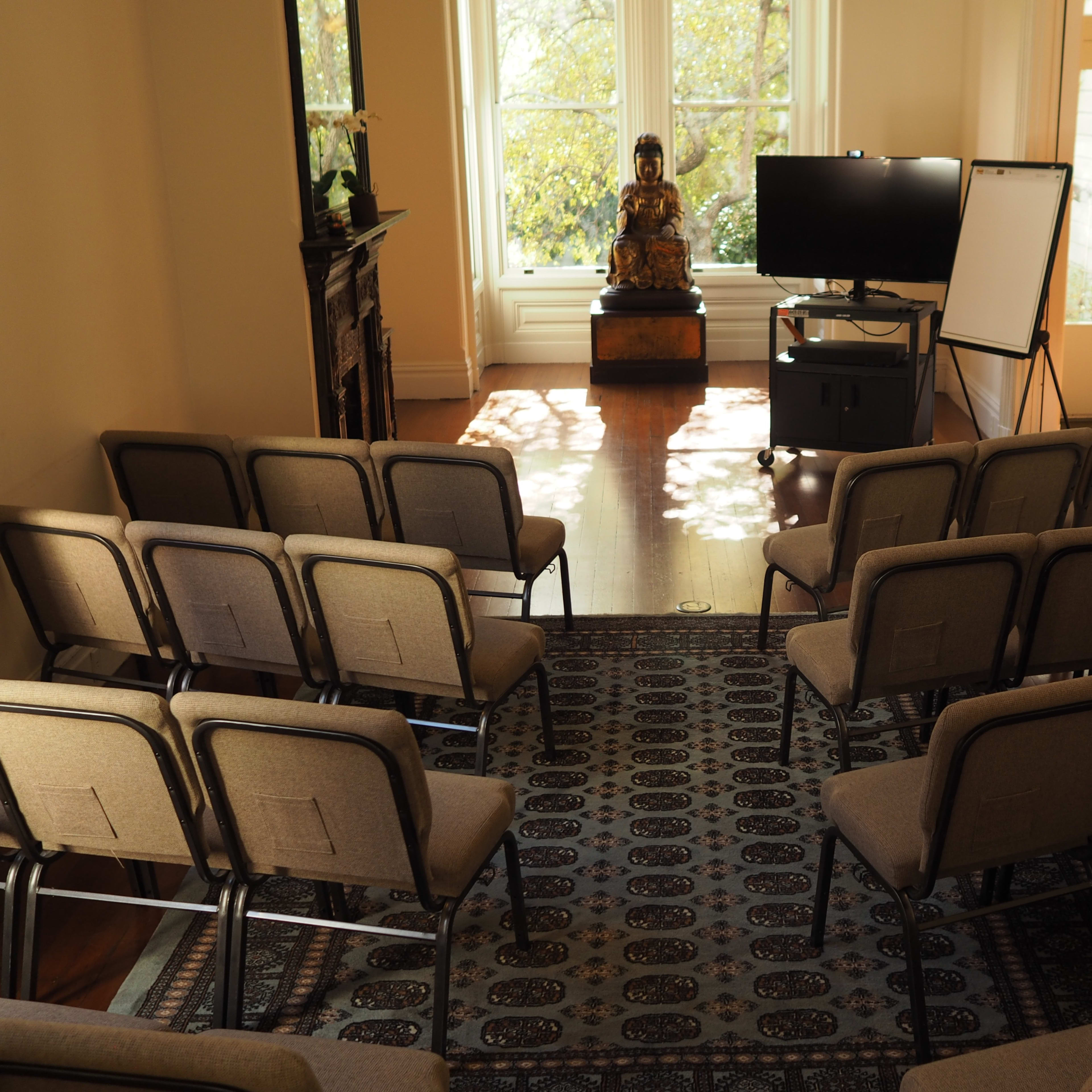 A small seminar room features rows of chairs facing a television and a whiteboard, with a statue of Buddha placed in the corner near a large window.