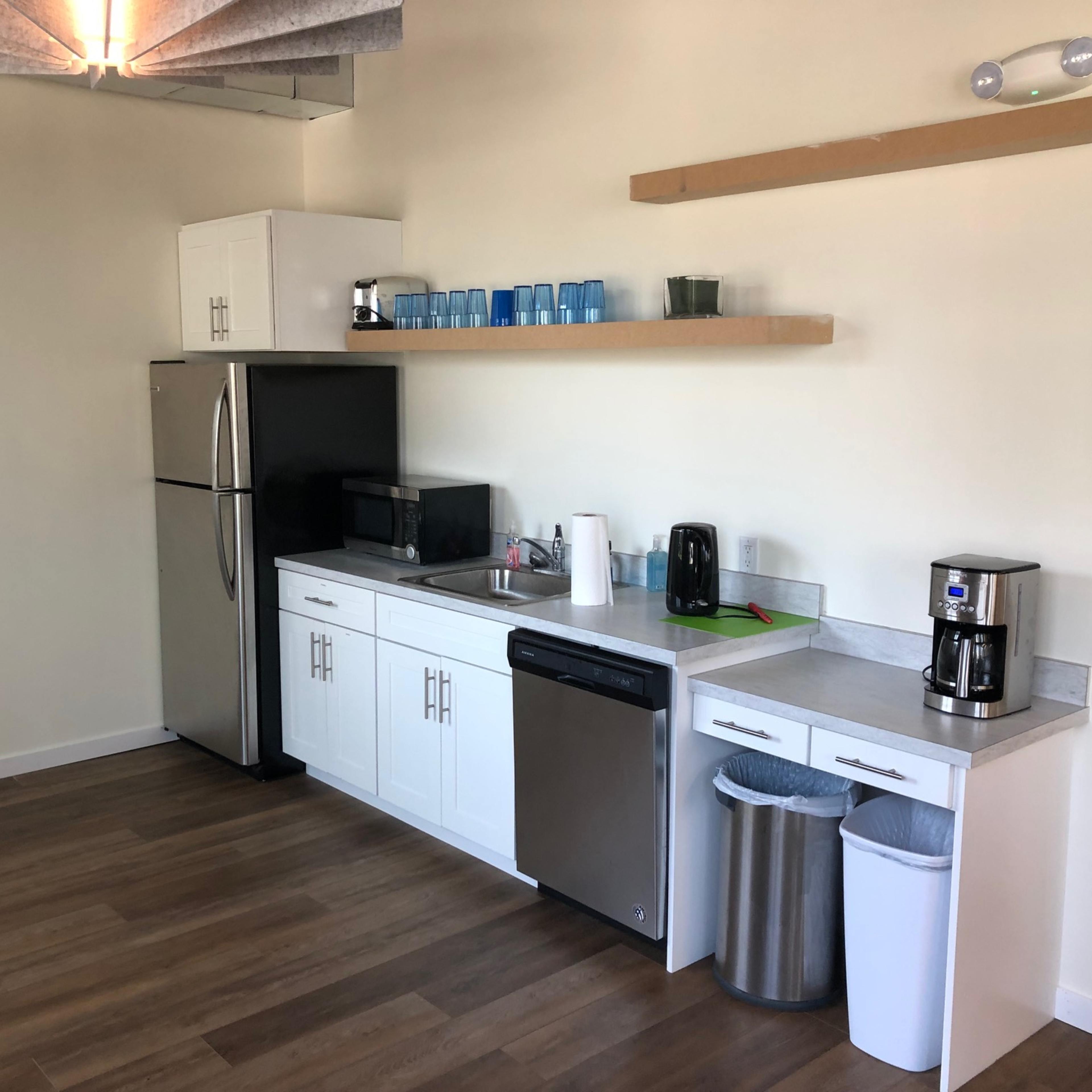 A modern kitchenette featuring a refrigerator, microwave, coffee maker, and a sink with white cabinetry and a wooden shelf above stocked with blue cups.