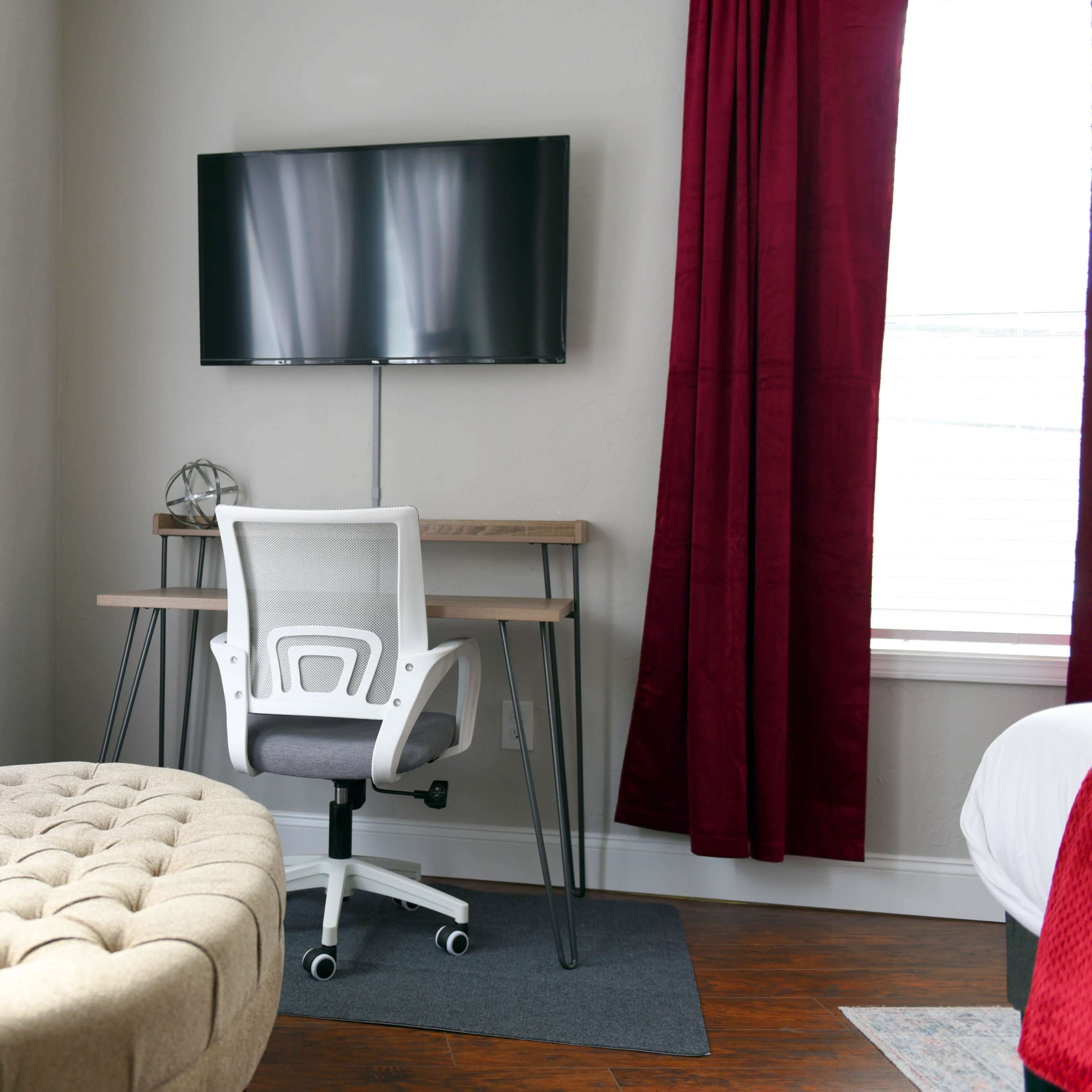 A modern workspace features a wooden desk with a white office chair, a wall-mounted TV, and red curtains, alongside a tufted beige ottoman on a hardwood floor.