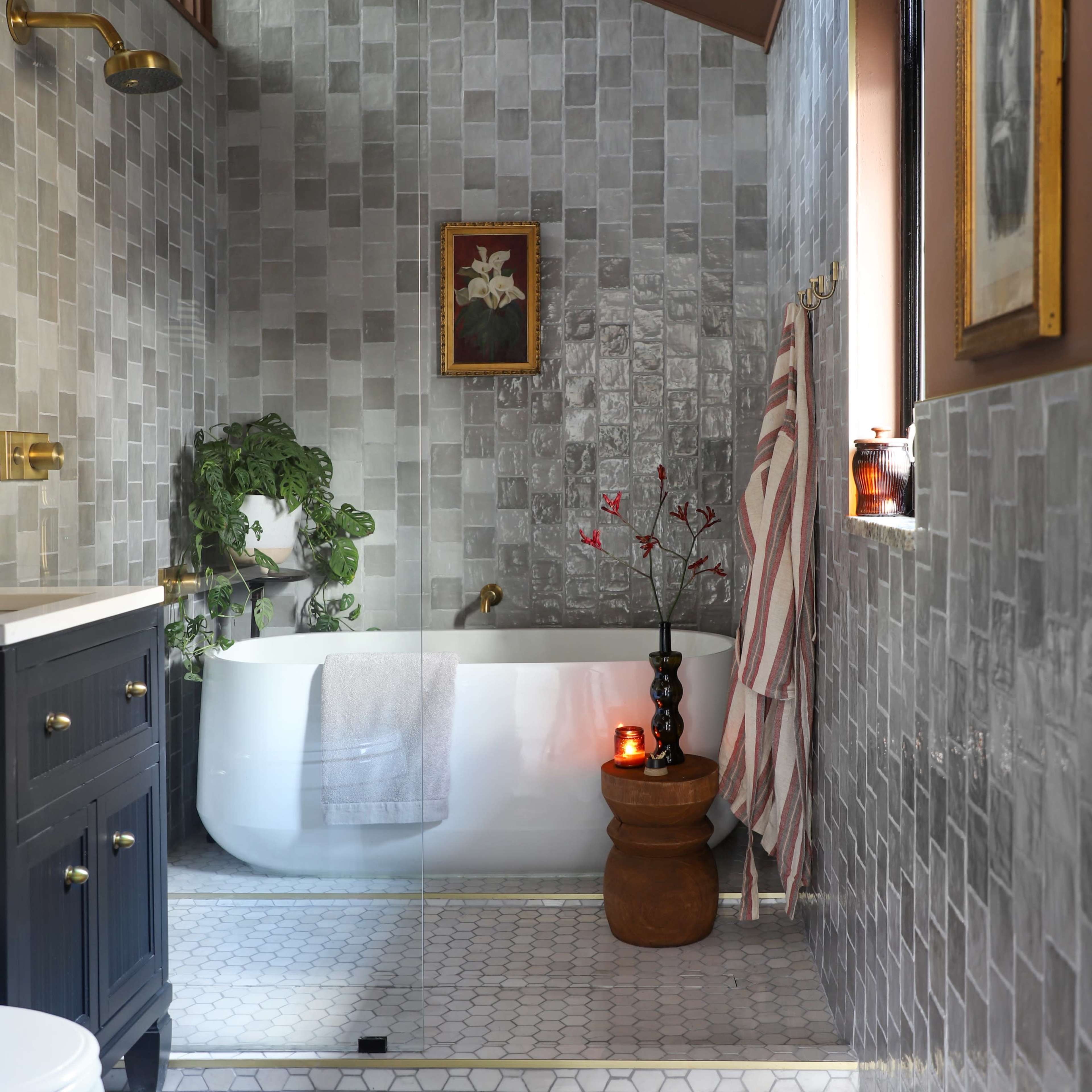 The bathroom features a freestanding tub, gray tiled walls, and a wooden cabinet with a small plant and decorative items.