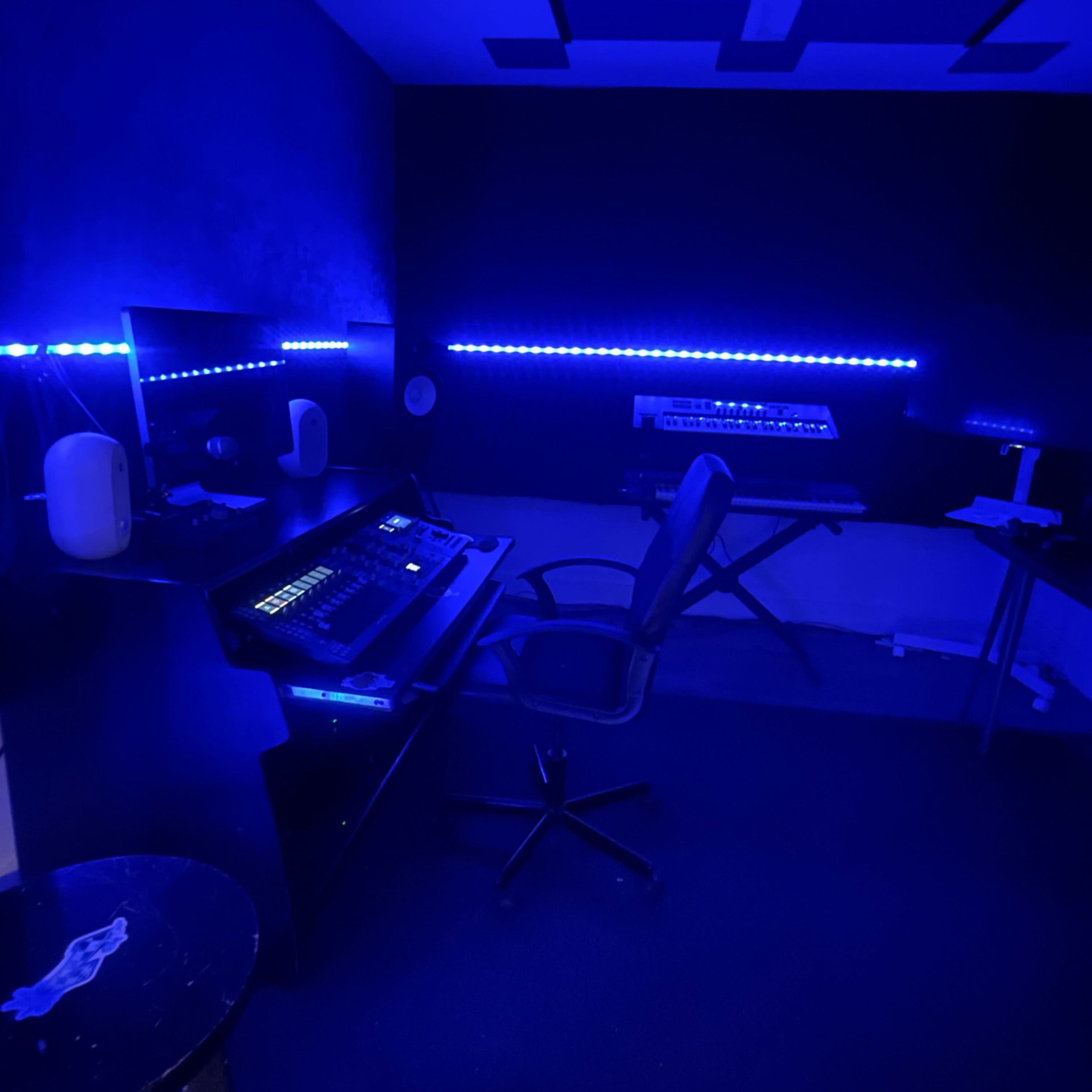 A dimly lit music studio with blue LED lighting, featuring a desk with equipment, a chair, and musical instruments.