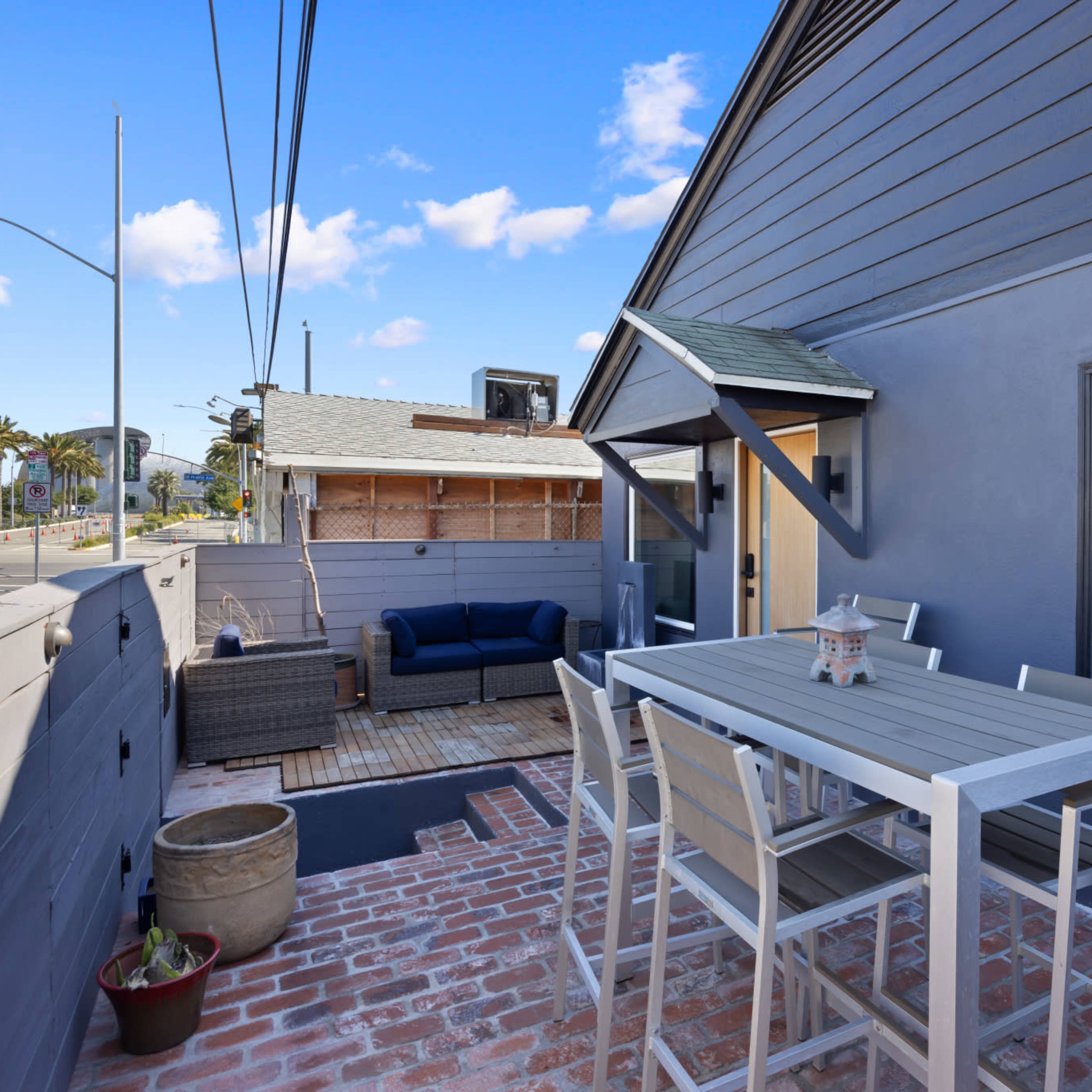 A patio area with a dining table and chairs, a couch in the background, and a view of a street and palm trees.