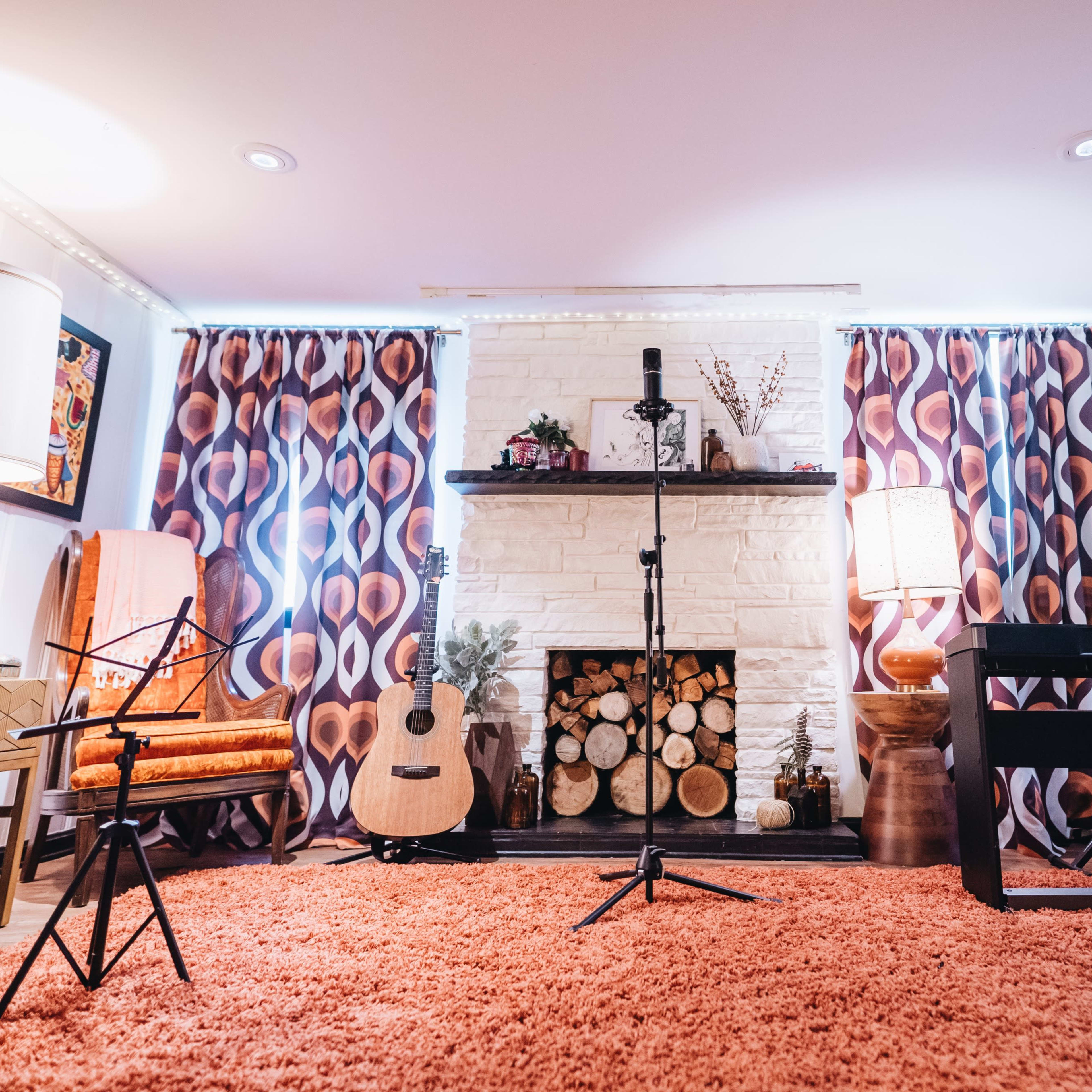 A cozy living room with a fireplace, decorated with guitars, a microphone on a stand, and two lamps, all set on an orange rug.