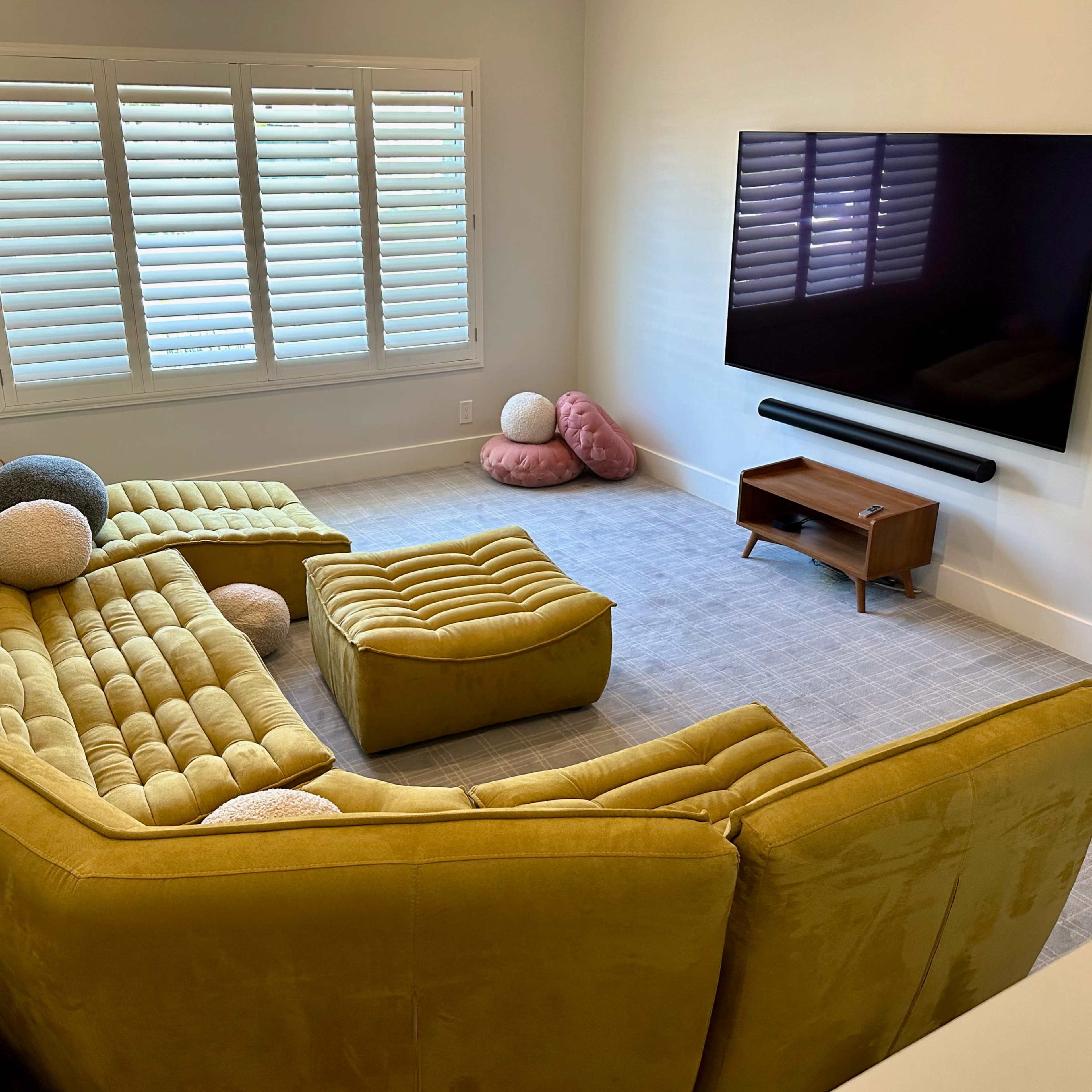 The room features a yellow sectional sofa, a matching ottoman, a wooden TV stand, and several decorative pillows on a light gray carpet.