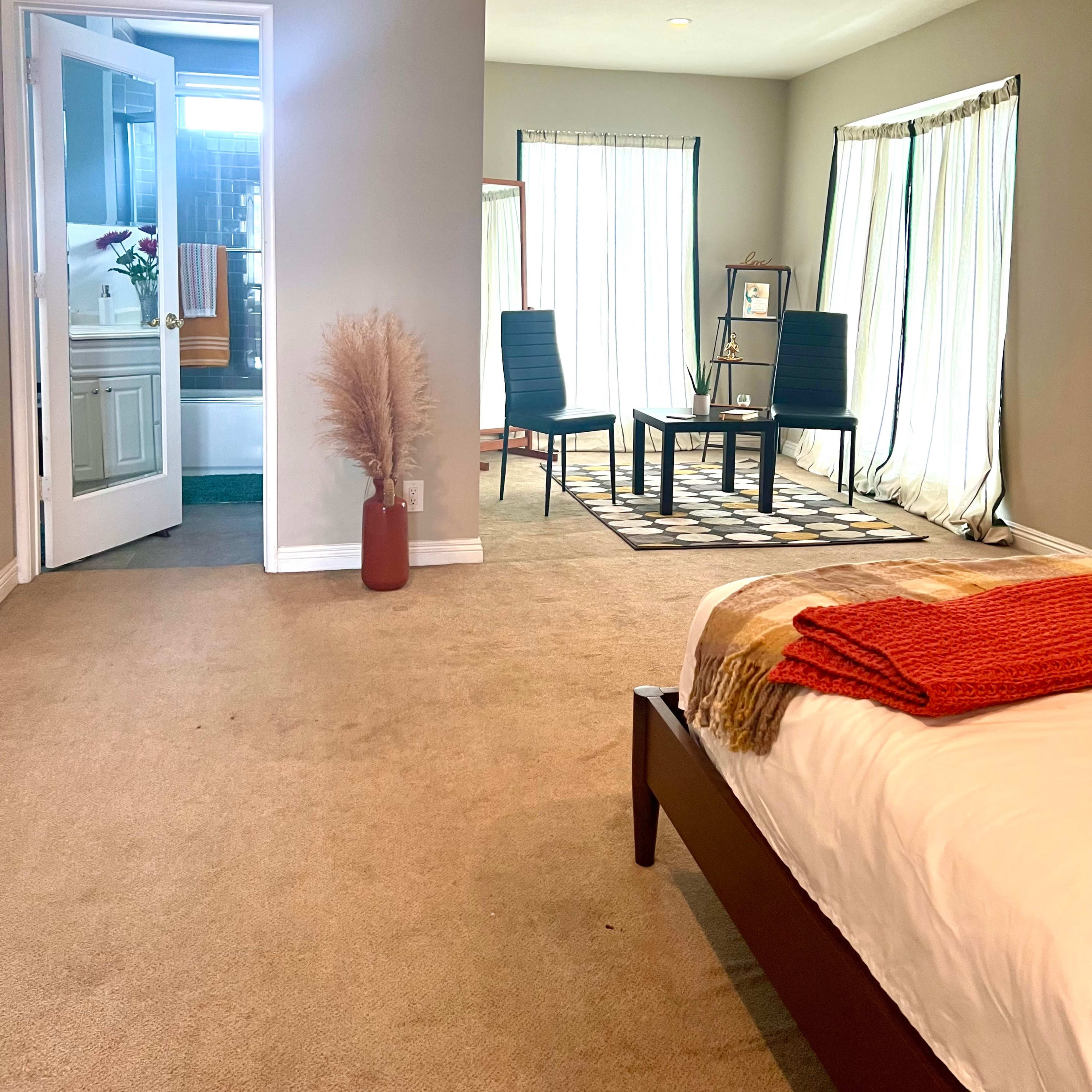 A spacious bedroom features a bed with an orange throw, a small sitting area with two chairs and a table, and doors leading to a bathroom.