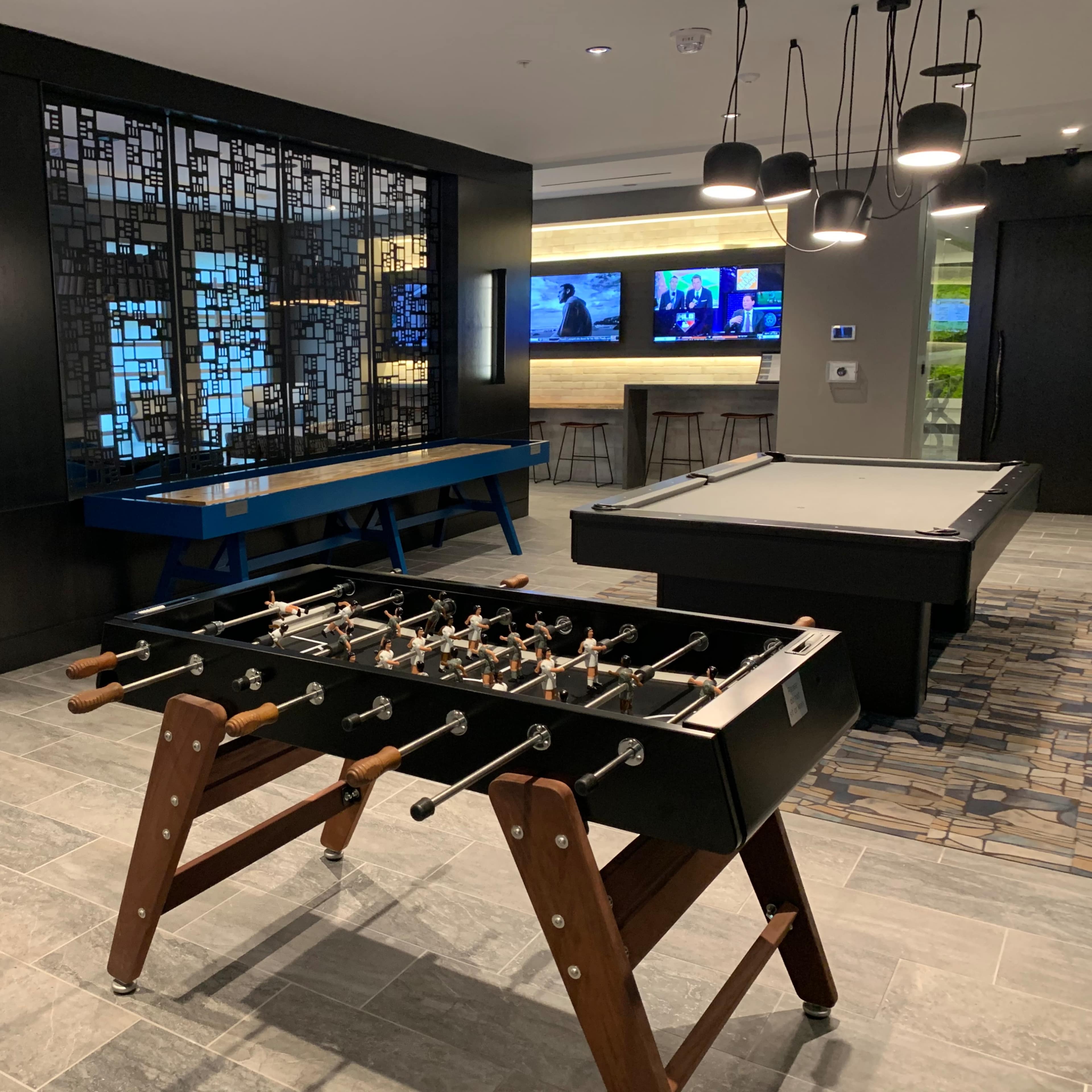 A recreational room featuring a foosball table in the foreground, a pool table in the background, and several screens mounted on the wall.
