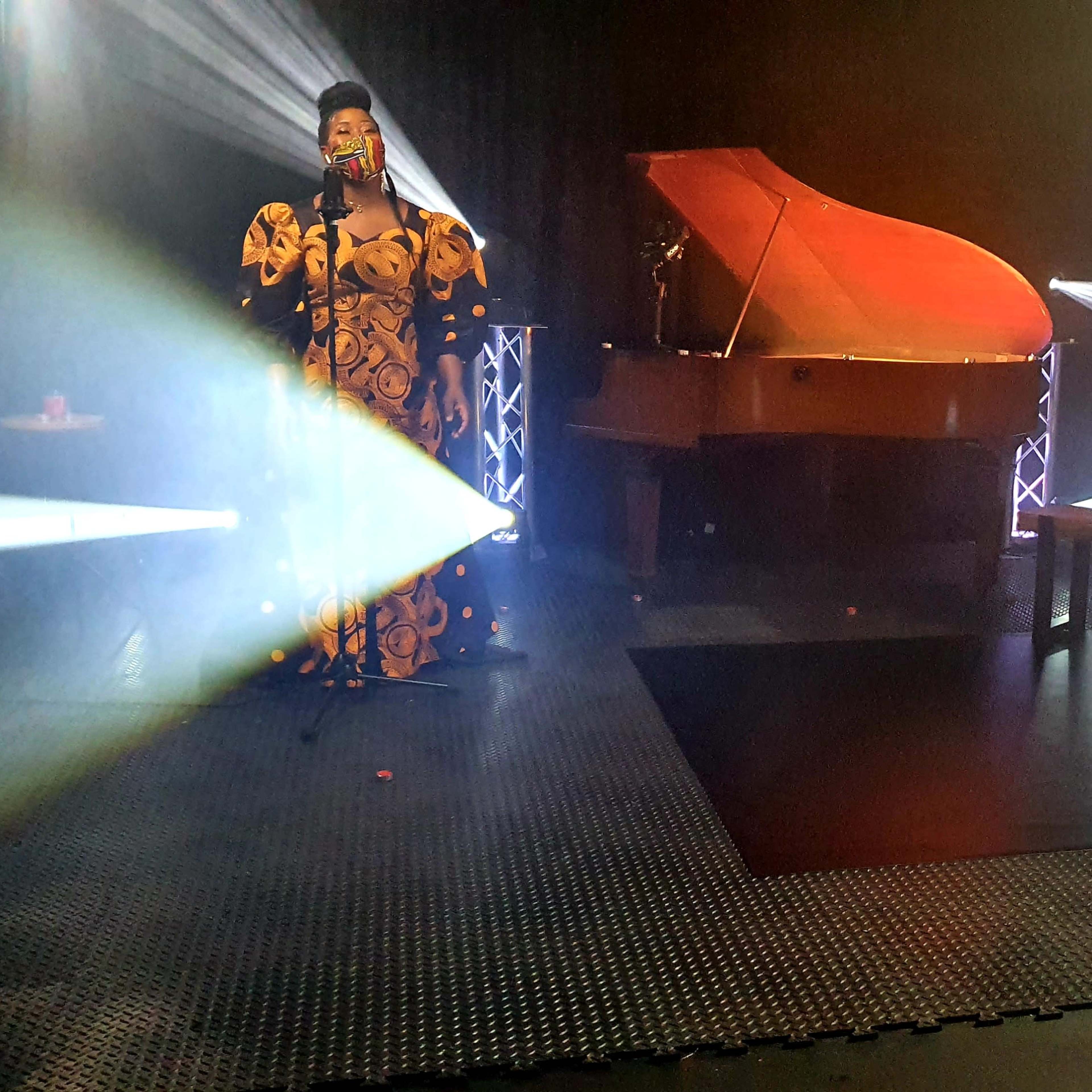 Three performers stand on a stage with a grand piano, shrouded in light beams and a misty atmosphere.