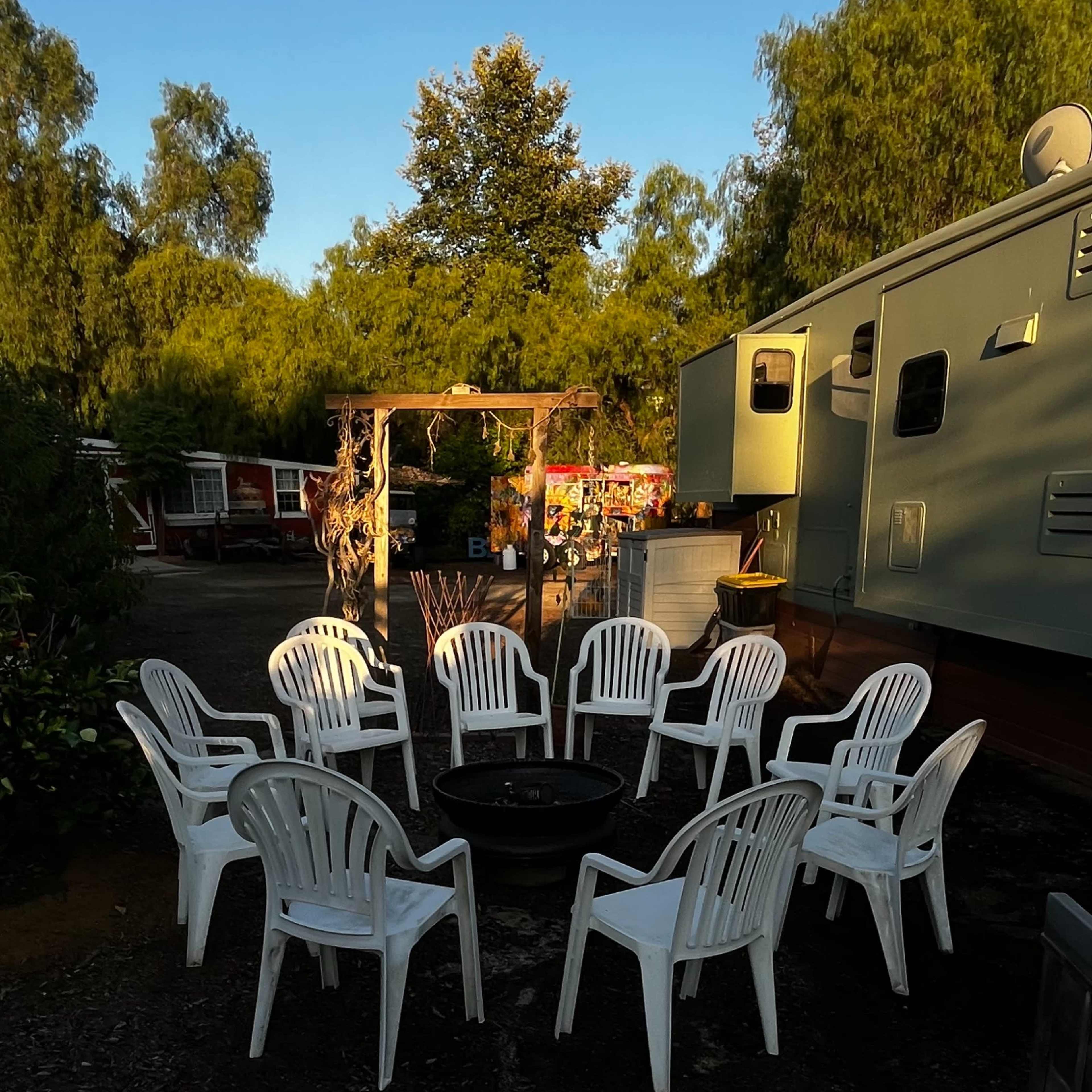 A circle of white plastic chairs surrounds a fire pit in an outdoor area next to a trailer, with trees in the background.