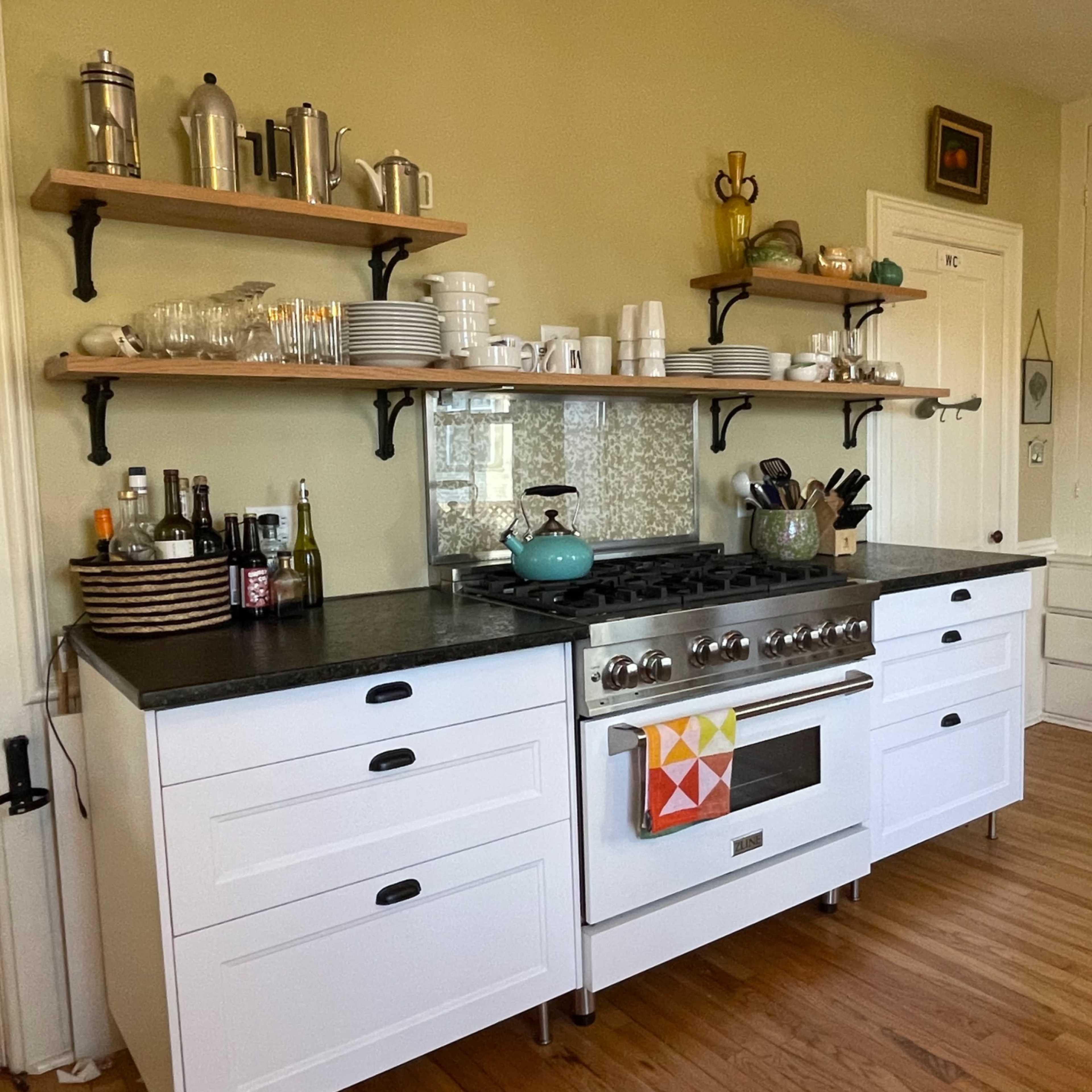 A kitchen with a white gas stove, black countertop, open shelves holding various kitchenware, and a wall of drawers in a light-colored room.
