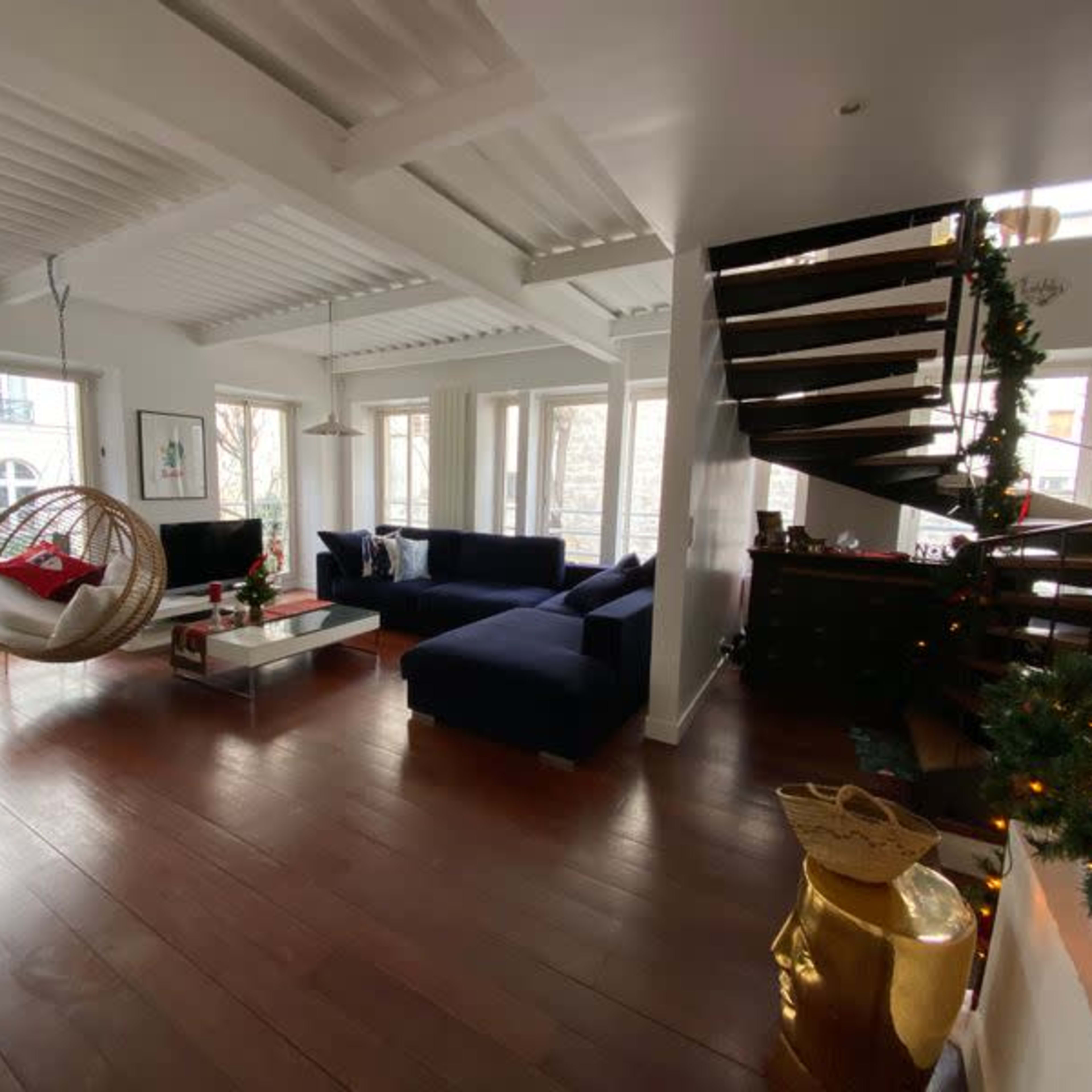 A modern living room features a large sectional couch, a hanging chair, and a staircase with wooden steps, surrounded by large windows that provide natural light.