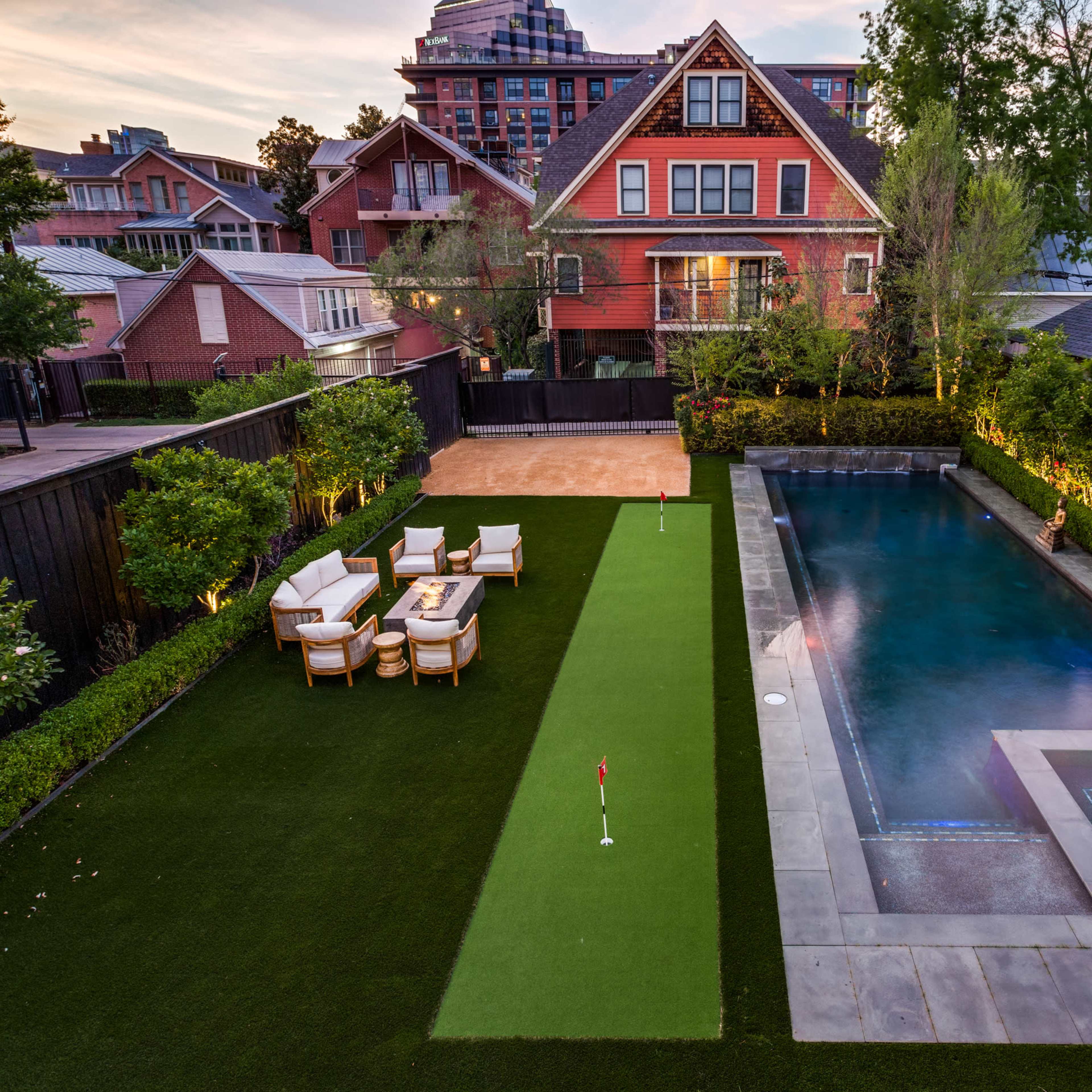 A well-maintained backyard featuring a green putting green and a rectangular swimming pool, bordered by manicured shrubs and a red-brick house in the background.