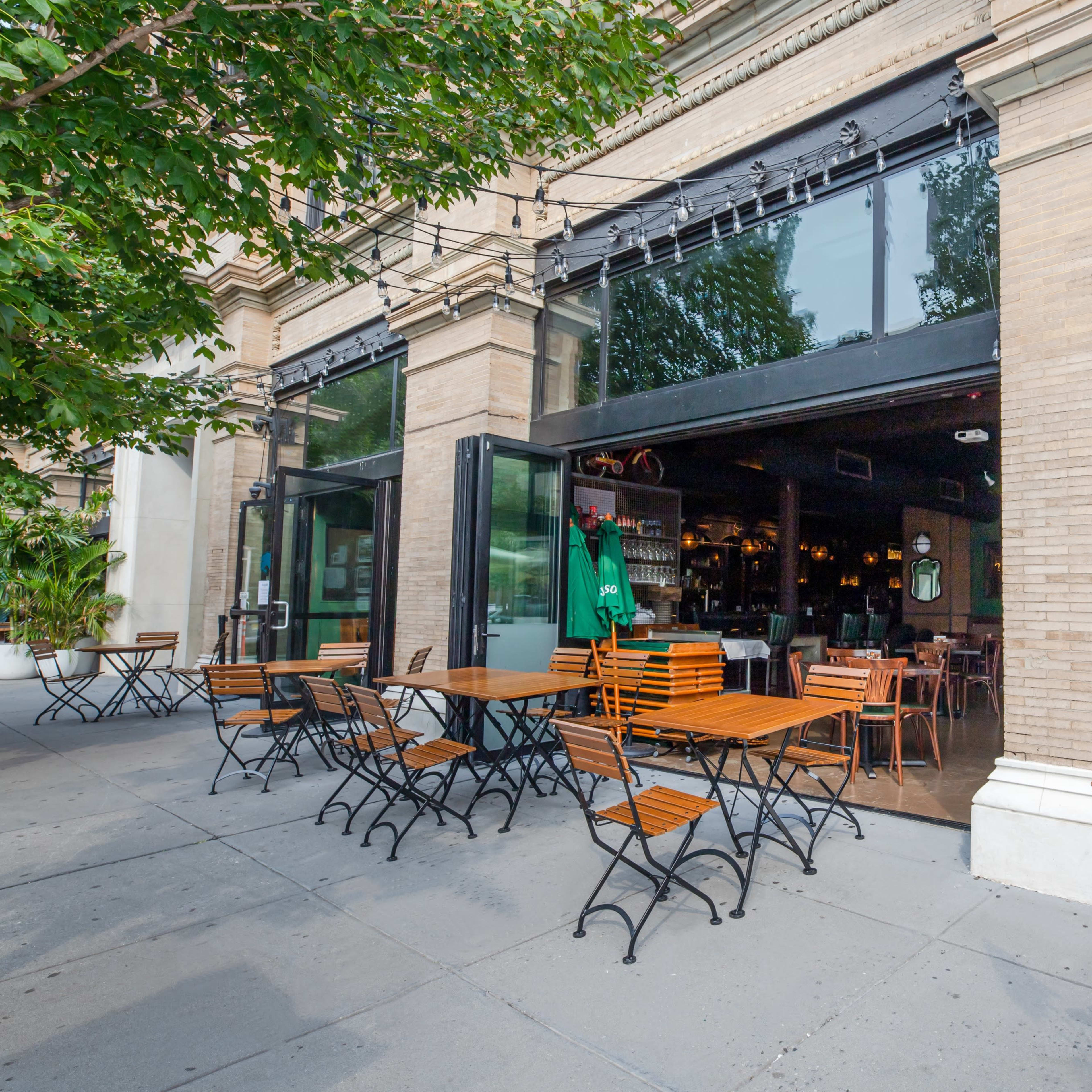 An outdoor seating area with wooden tables and chairs is situated in front of a restaurant with large open windows.